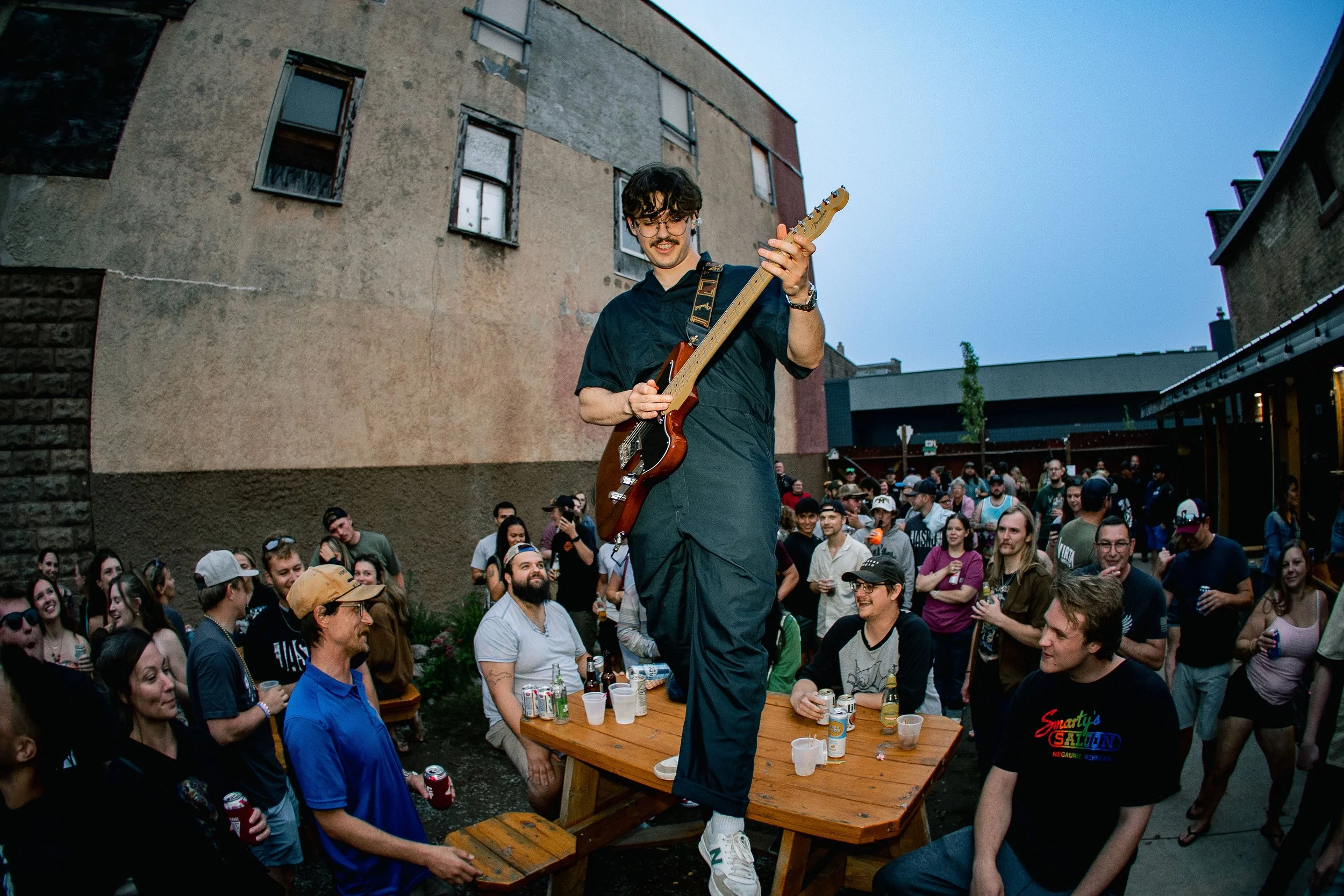 A man with glasses and a mustache playing an electric guitar while standing on a table at an outdoor party. He is surrounded by a crowd of people watching and smiling, with some holding drinks, during dusk or early evening.