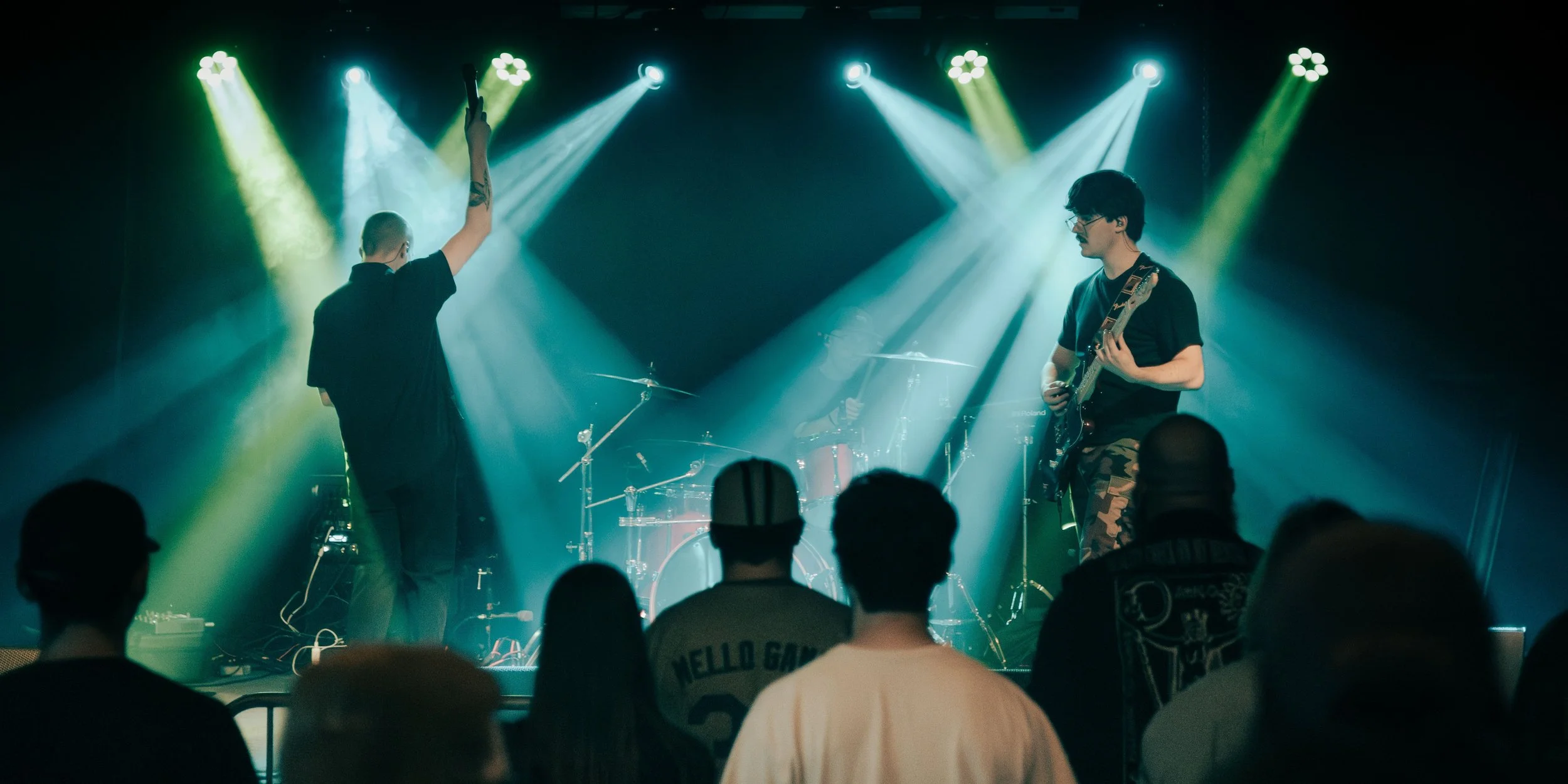A band performing live on stage with three musicians and an audience watching, illuminated by green and white stage lights.