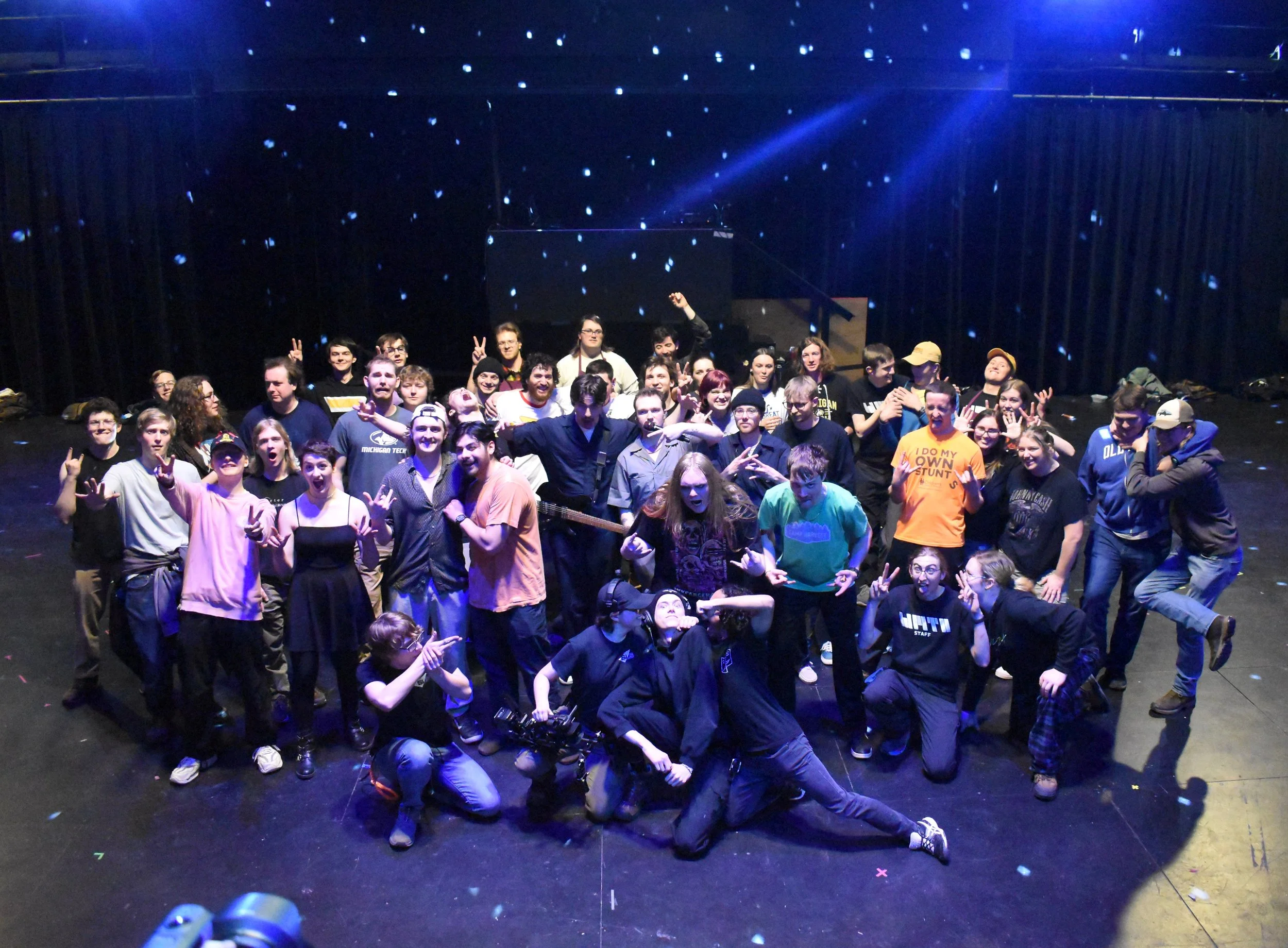 Group of people gathered on stage for a photo, some making peace signs or other gestures, with a dark backdrop and stage lighting.