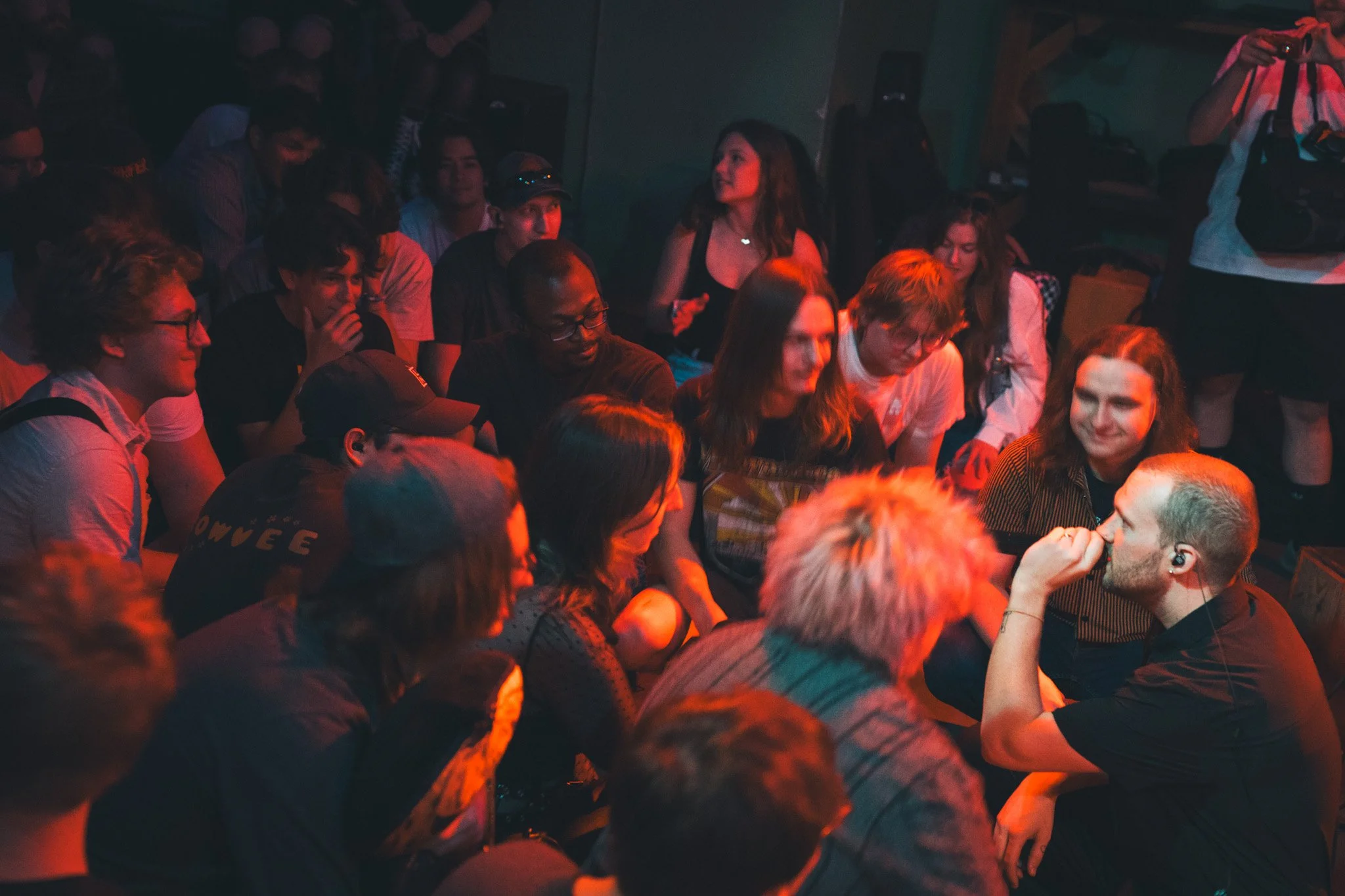 A group of people sitting in a dark room, engaged in a conversation or performance, with some smiling and others focused, illuminated by red lighting.