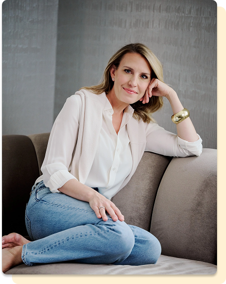 A woman sitting on a couch in a bright, casual outfit, resting her head on her hand and smiling.