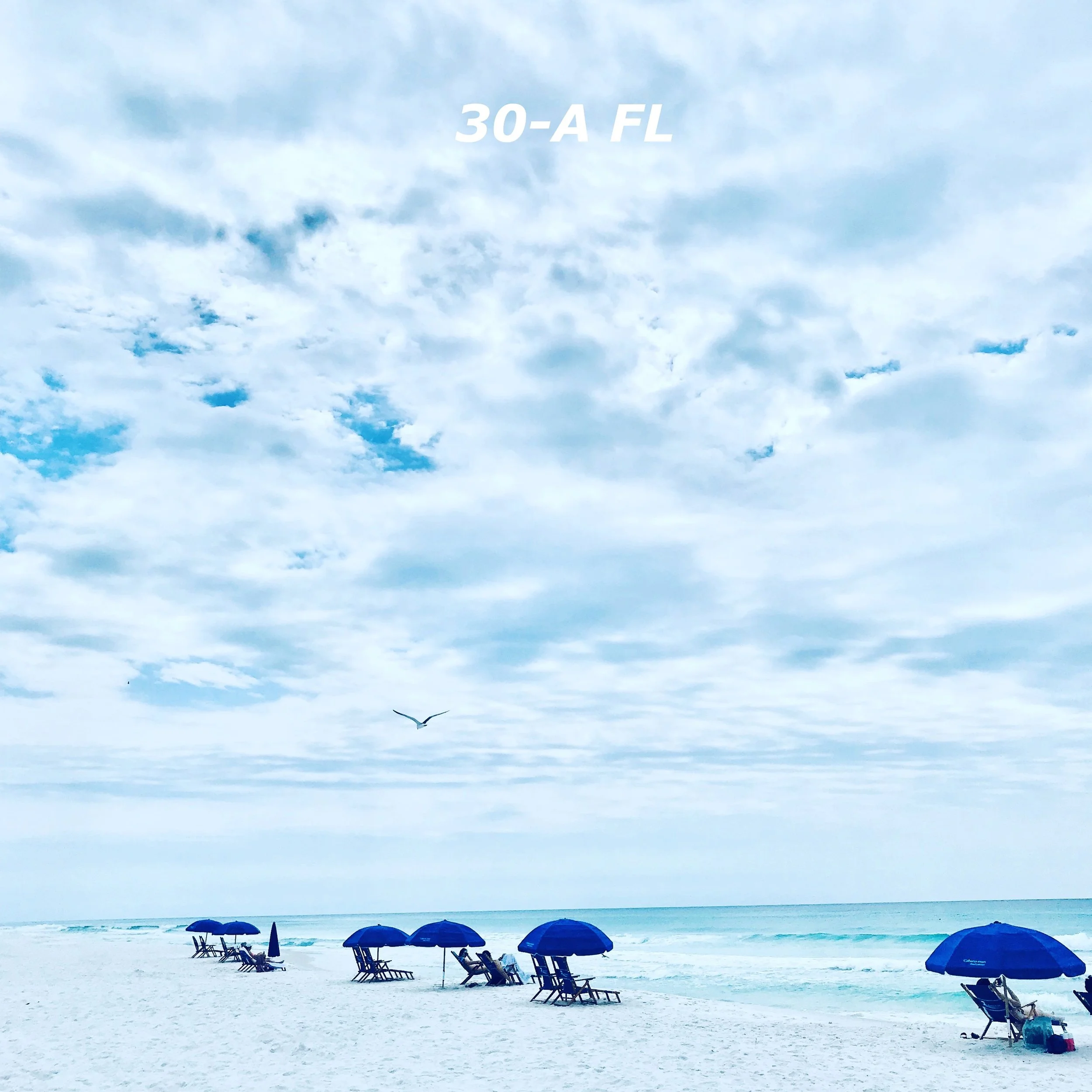 Beach scene with seven blue umbrellas, empty chairs, and a few people sitting on the chairs, along with a bird flying in a cloudy sky. The sand is white and the ocean is calm in the background.
