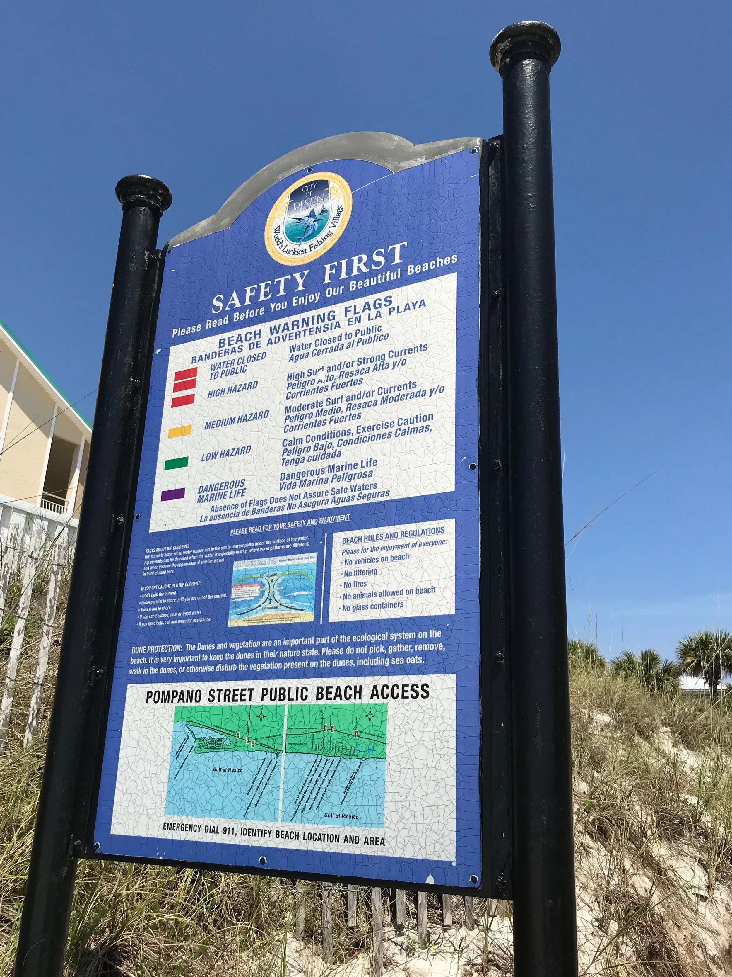 Beach safety sign at Pompano Street Public Beach Access in Miramar Beach, listing warning flags, beach rules, and regulations, with a map of the beach access area and instructions for emergency contact.