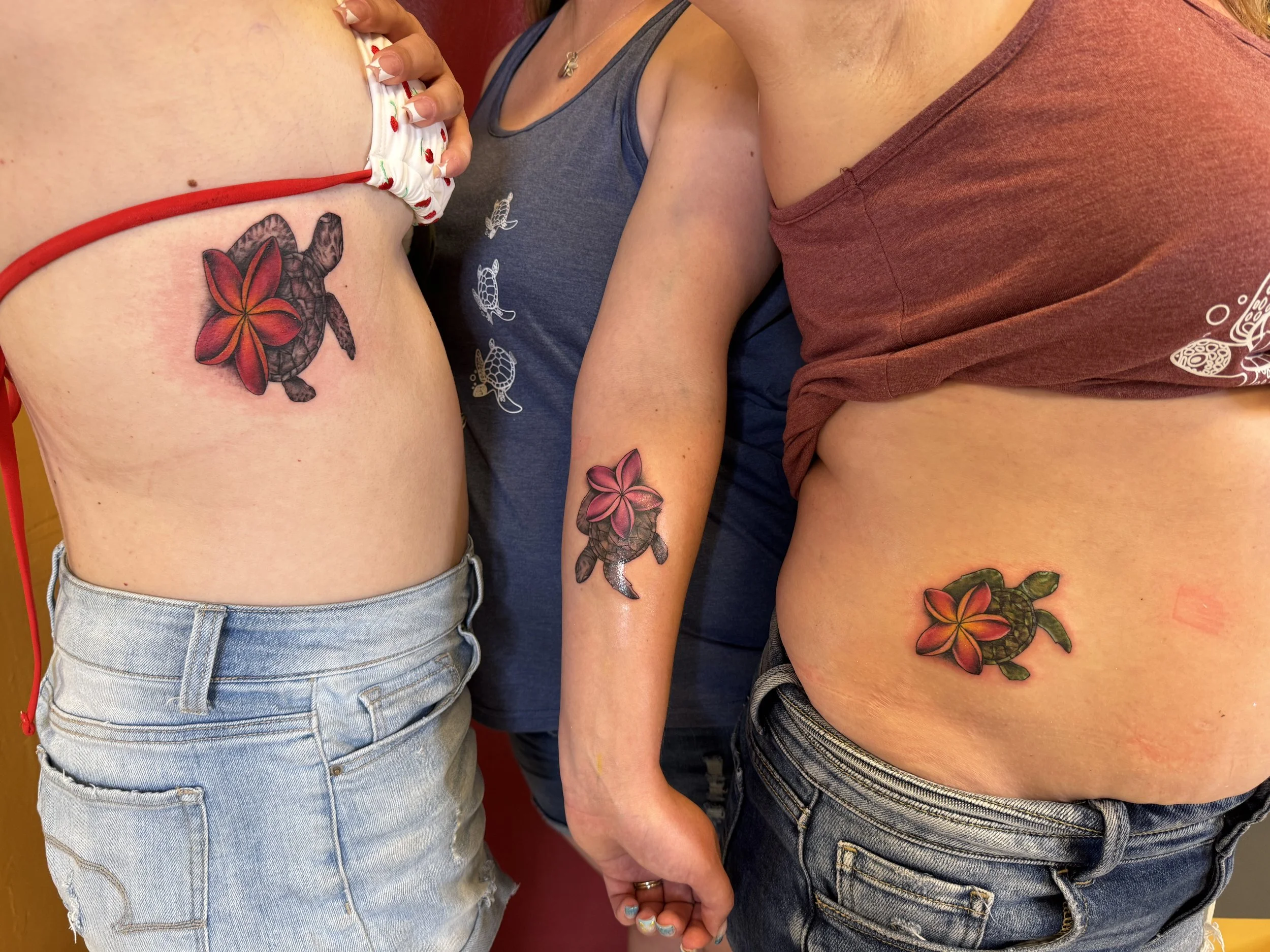 Three people standing close together, holding hands, with matching turtle hone plumeria flower tattoos on their arms and colorful flower tattoos on their hips.