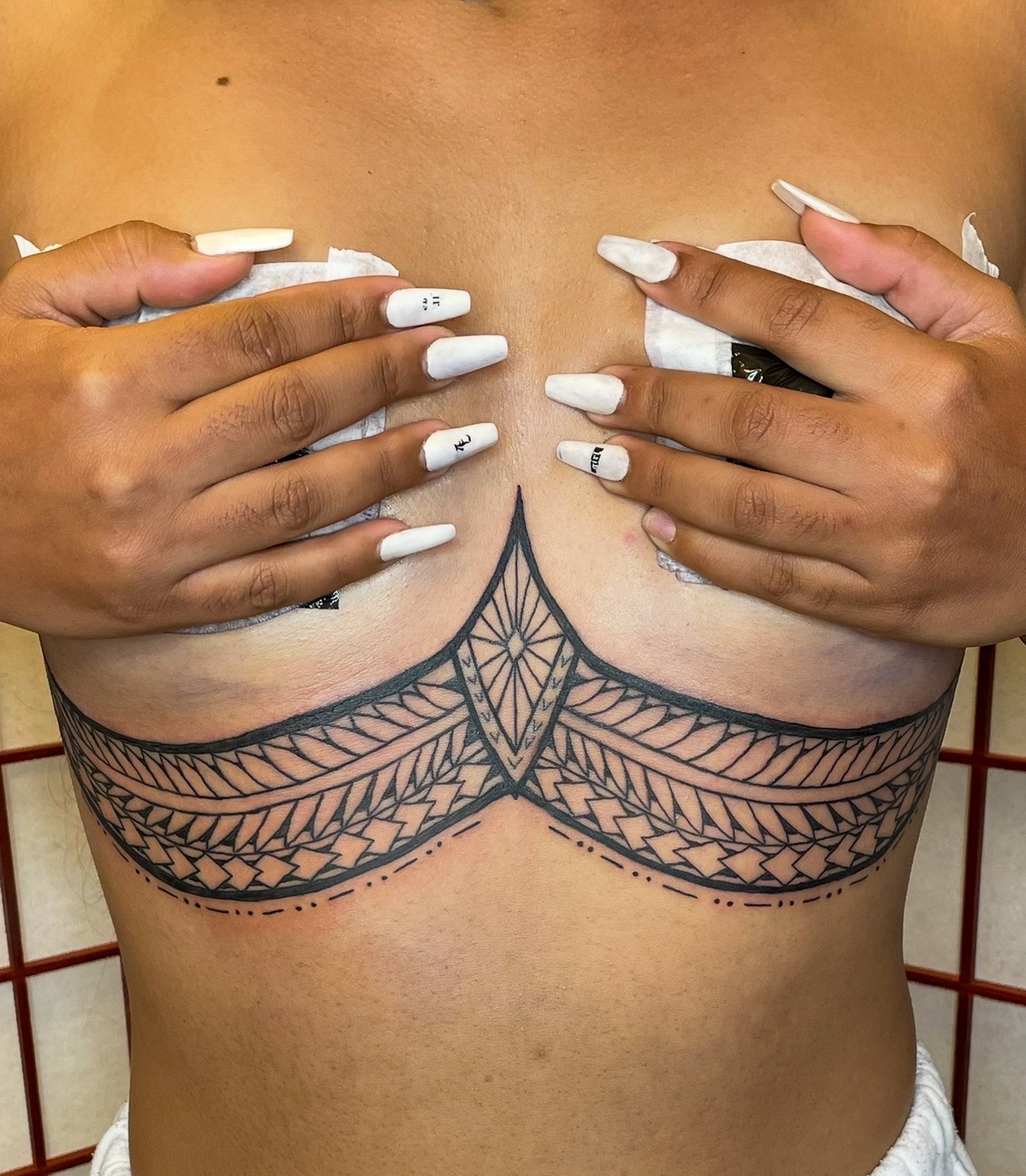 Close-up of a person's chest with decorative black tattoo of geometric design, while holding white tissue paper near their breasts with nails painted in white and black text.