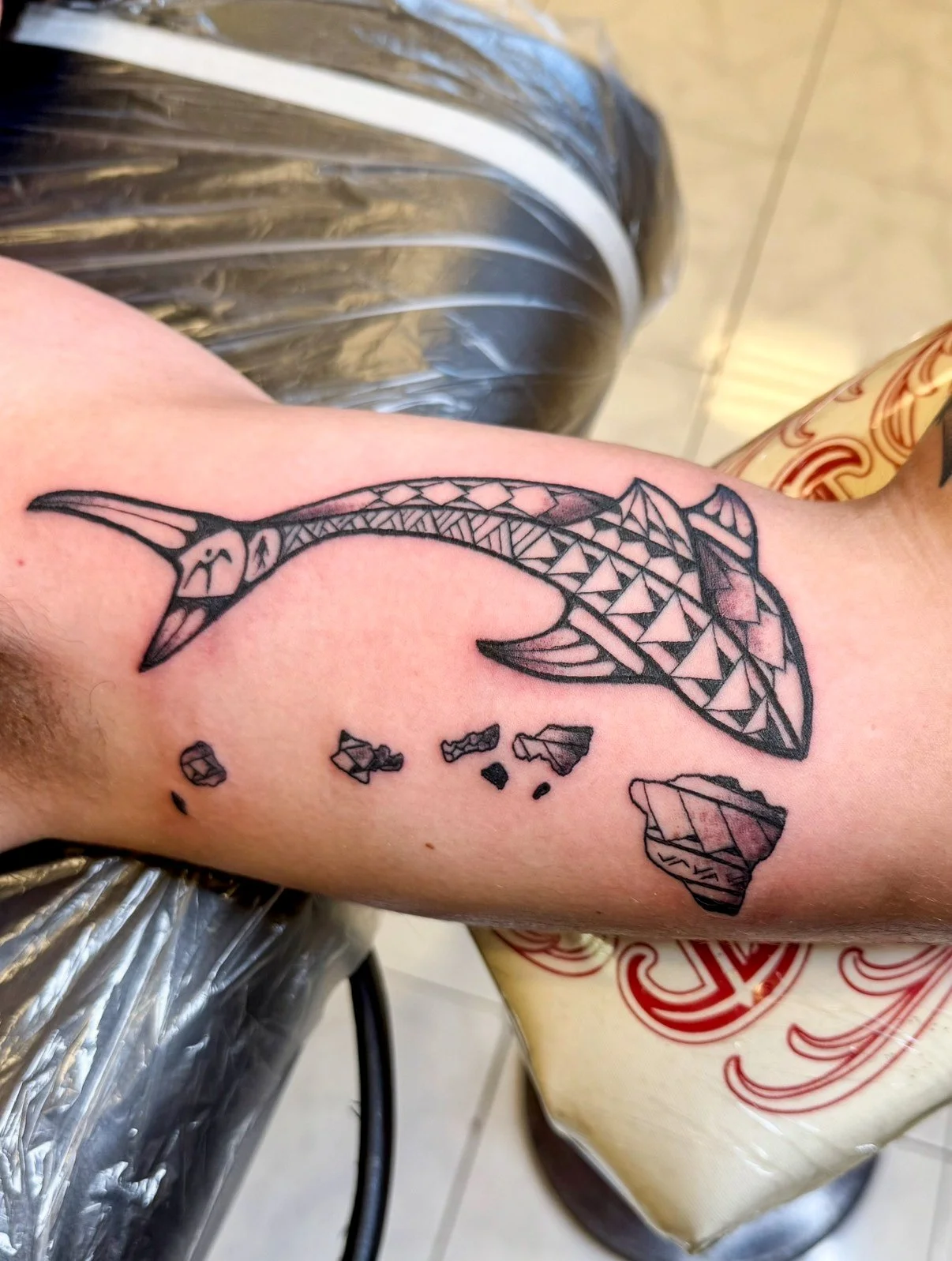 Tattoo of a stylized whale with geometric patterns on the person's arm, with a few smaller broken pieces below.