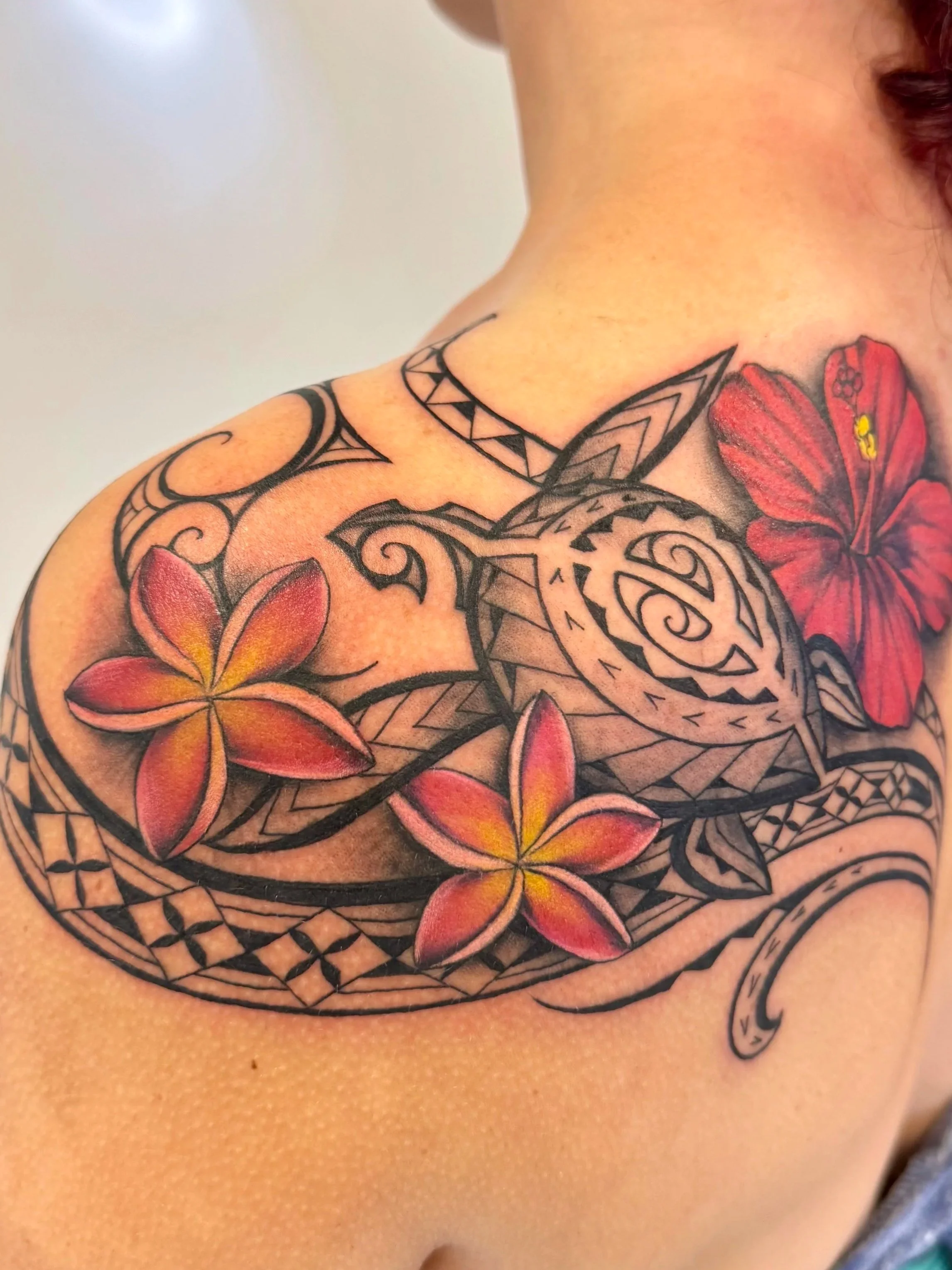 Polynesian sea turtle tattoo on shoulder and upper back featuring Hawaiian tribal patterns, plumeria flowers, hibiscus flower, and black linework with red and yellow color accents, custom Polynesian floral tattoo by Monarch Tattoo Hawaii on Oahu