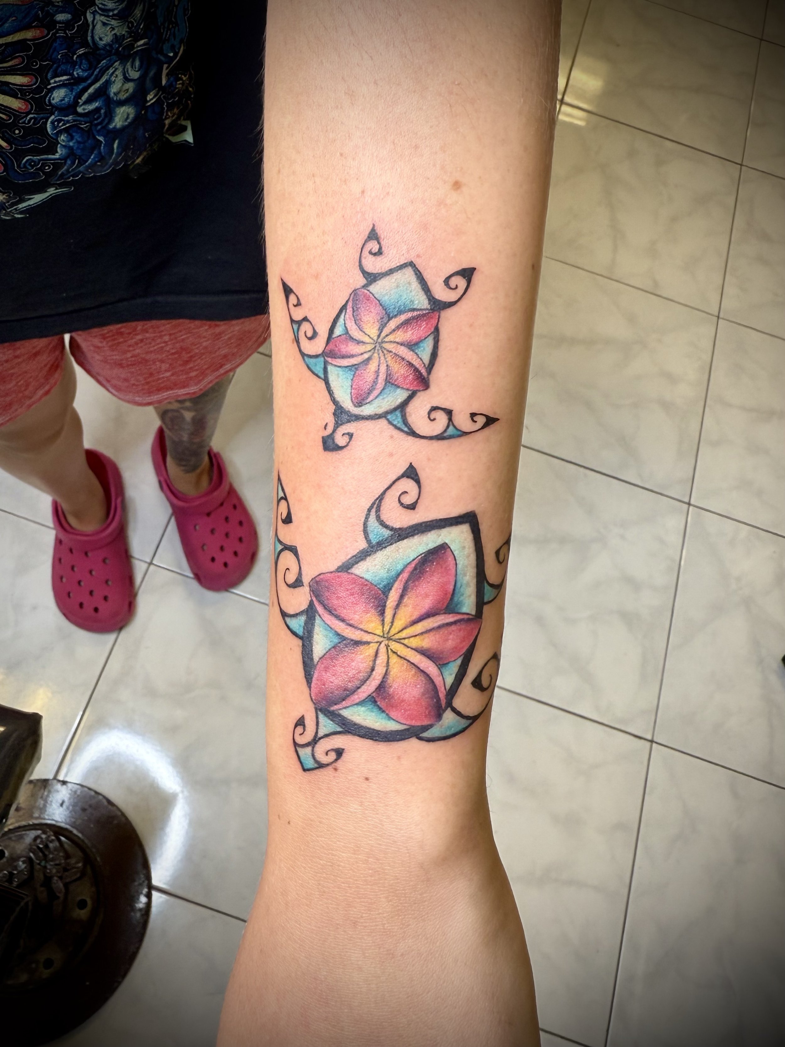 Colorful tattoo of pink and orange plumeria flowers on a person's forearm, with decorative black swirls surrounding the flowers.