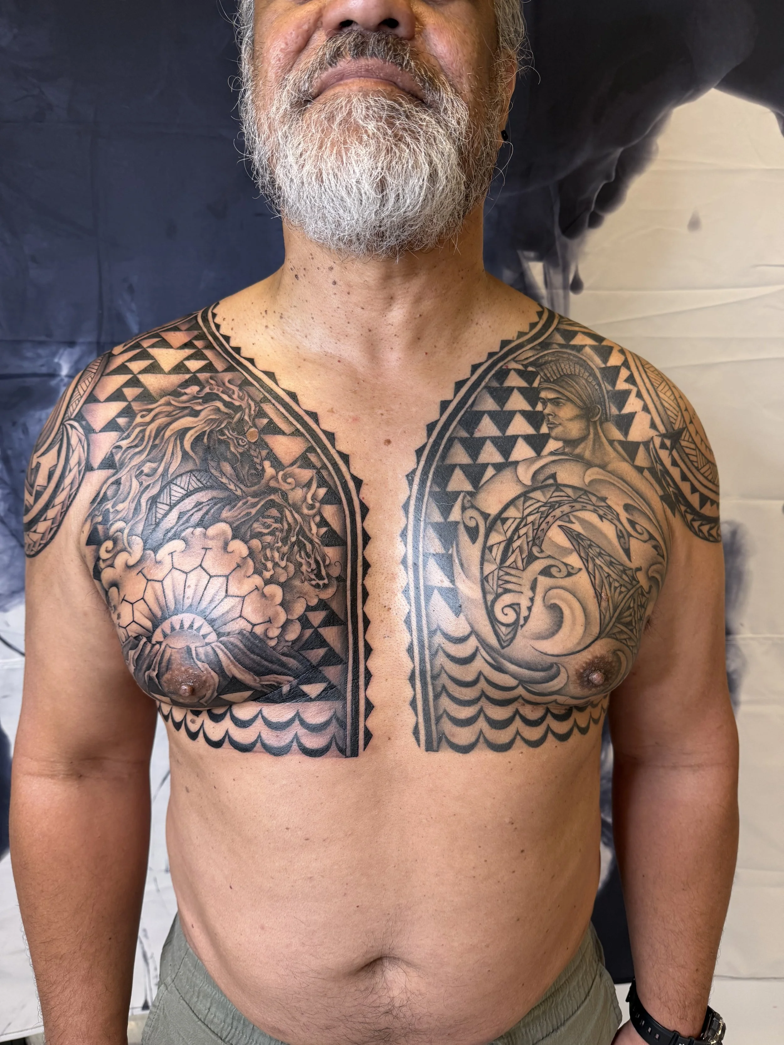 Upper body of a man with a bald head, gray beard, and extensive black ink Polynesian tattoos covering his chest and shoulders, featuring a lion and a traditional warrior figure, against a dark background.