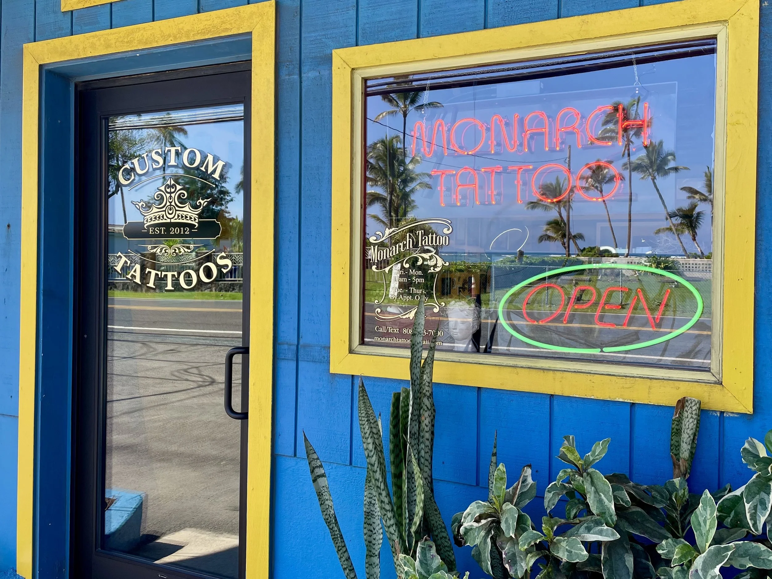 Monarch Tattoo Hawaii studio exterior featuring blue and yellow storefront, neon Monarch Tattoo sign, open window display, custom tattoo shop door signage, and tropical plants in a Hawaiian beach street setting