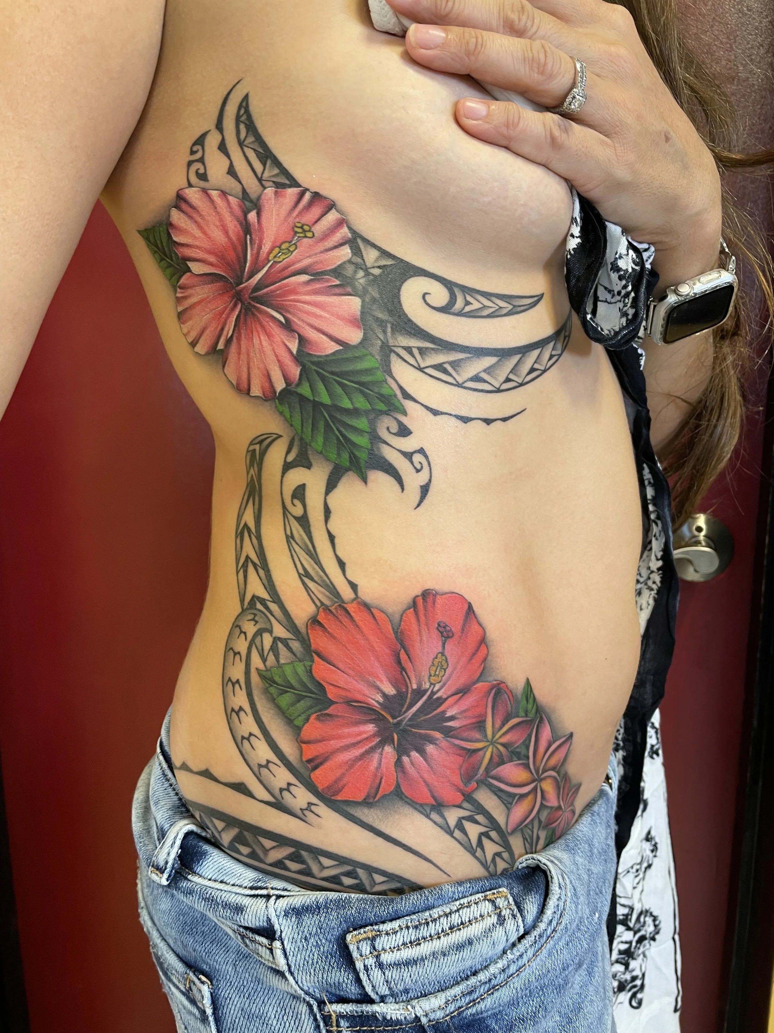 Colorful coverup tattoo of red hibiscus flowers, green leaves, and Polynesian tribal patterns on the side of a woman's torso.