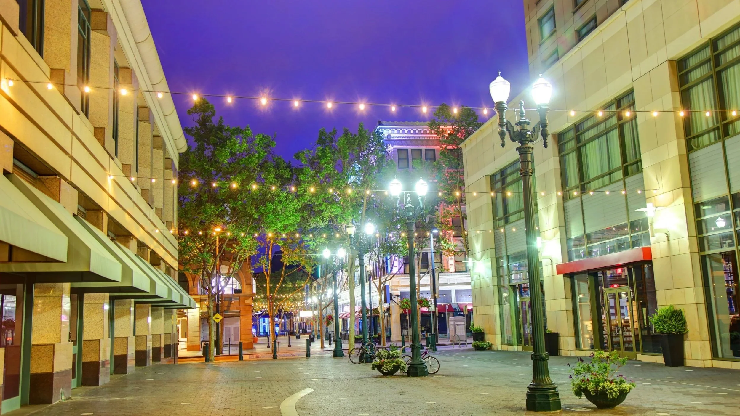 Empty city street at night with lit street lamps, trees, bicycles, buildings, and string lights overhead.