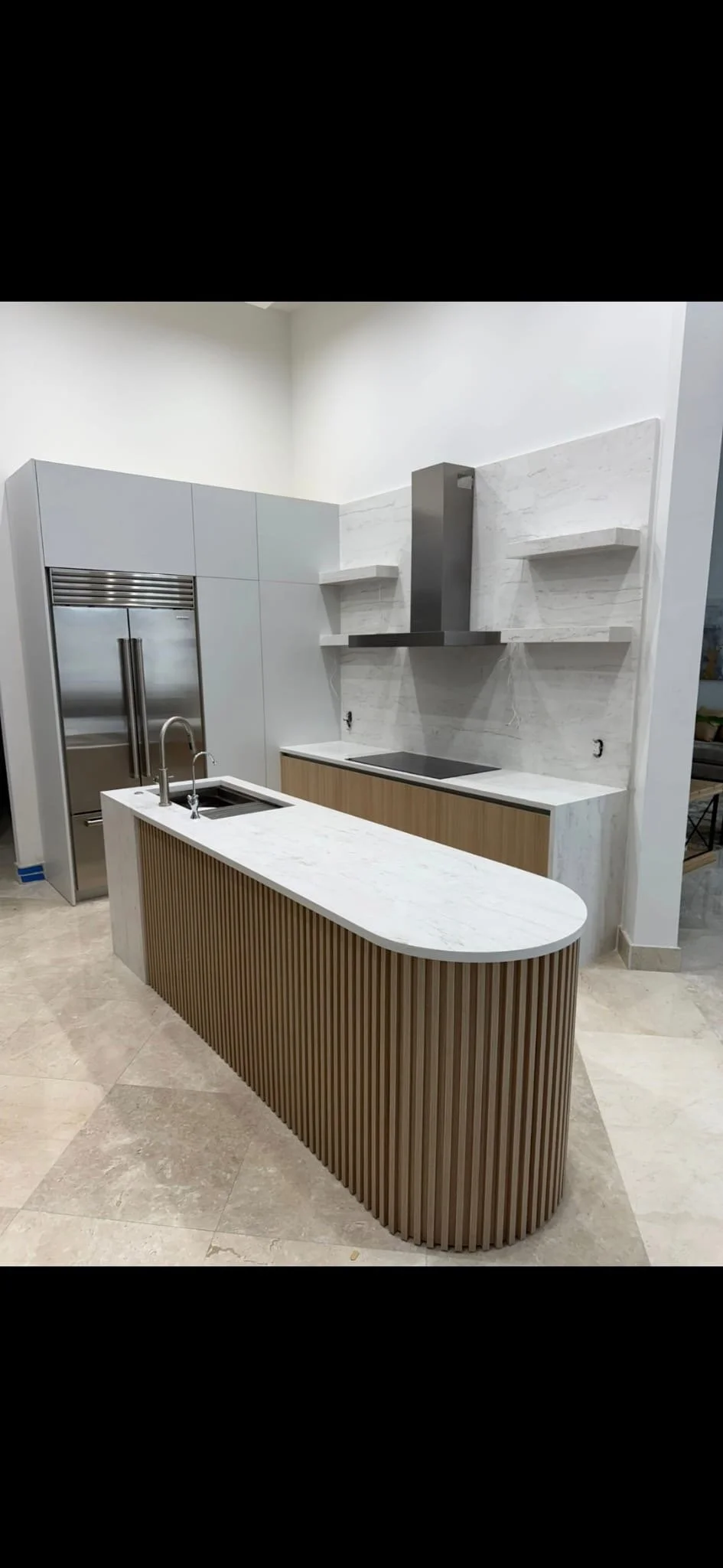 Modern kitchen with a curved island featuring a white marble top and vertical wooden slats, stainless steel refrigerator, black cooktop, stainless steel range hood, and open shelves on a white marble backsplash.