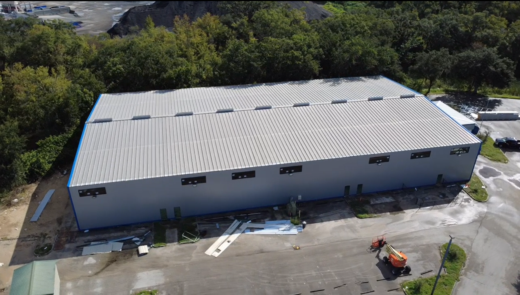 Aerial view of a large industrial building with a metal roof surrounded by trees, parking lot, and construction equipment.