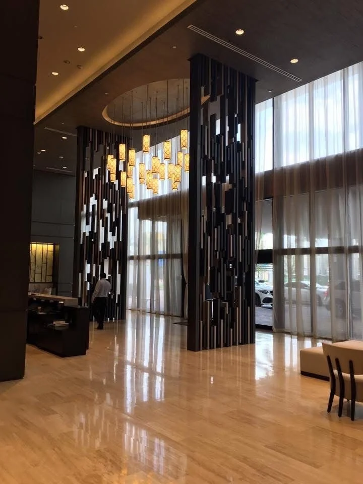 Elegant hotel lobby with tall decorative wooden partitions, a modern chandelier, large windows with sheer curtains, and a polished wooden floor.