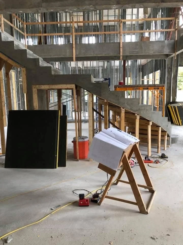 Construction site inside a building with an unfinished staircase and framing. Building materials, tools, and notes are present on a stand in the foreground.