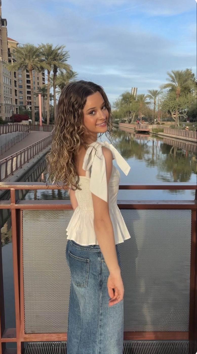 A young woman with curly brown hair smiling at the camera, standing on a bridge over a canal with palm trees and modern buildings in the background during the daytime.