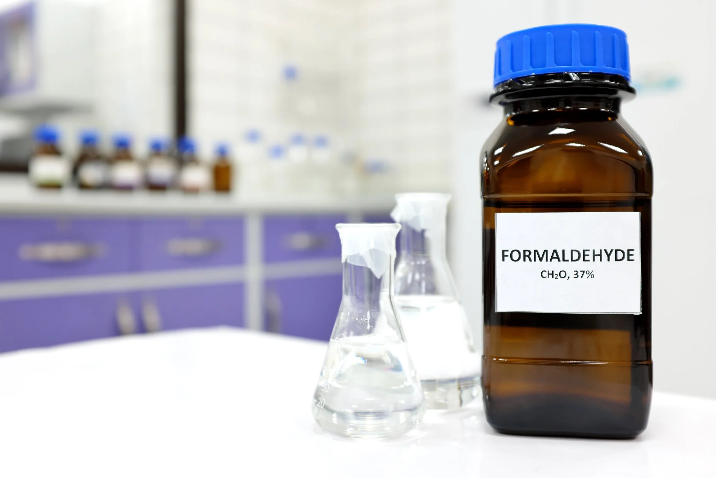 A brown bottle labeled 'Formaldehyde' with a blue cap, and two glass flasks filled with clear liquid, placed on a white surface in a laboratory setting.