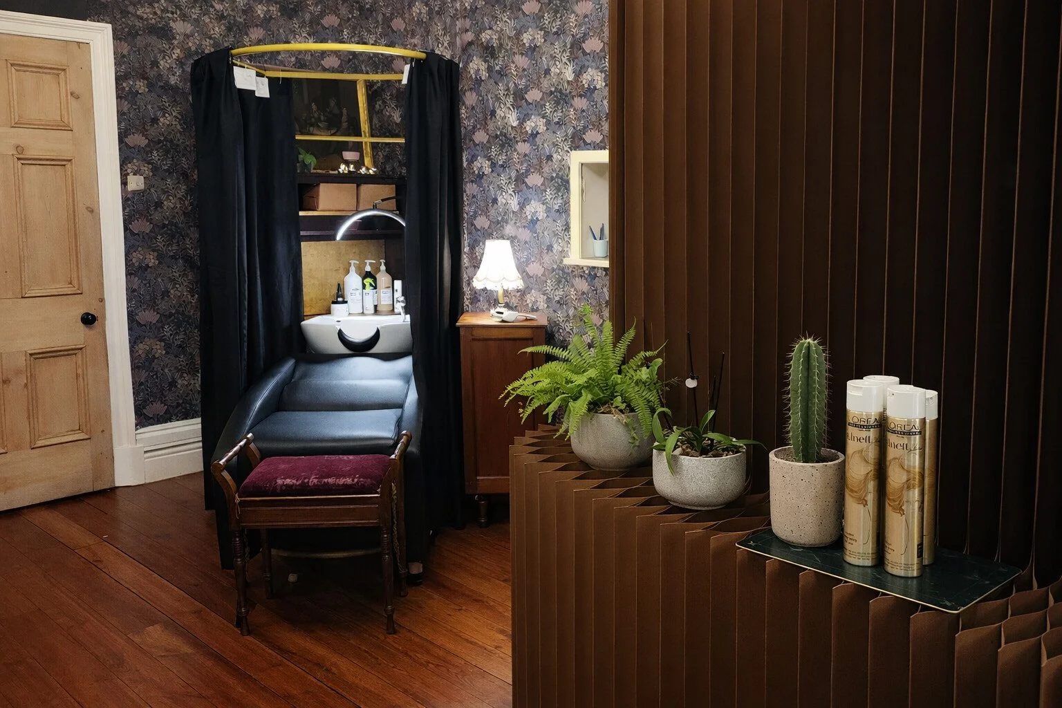 Interior room with wooden floors, a small sink with shampoo and bottles, a mirror, a lamp, plants, and a black curtain surrounding the sink area.