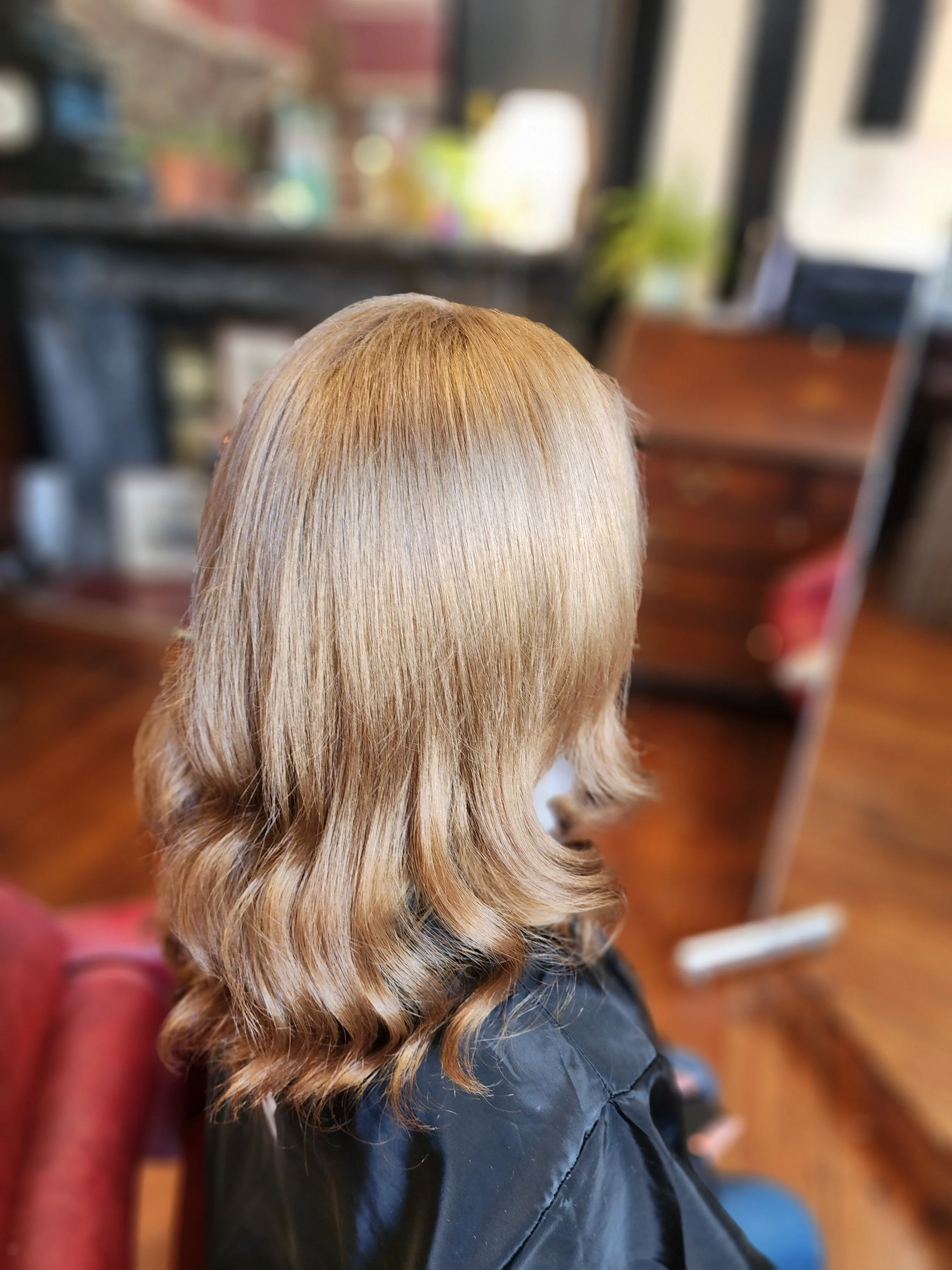 Close-up of a person with long, wavy, blonde hair, wearing a dark jacket, sitting in a salon with blurred background.