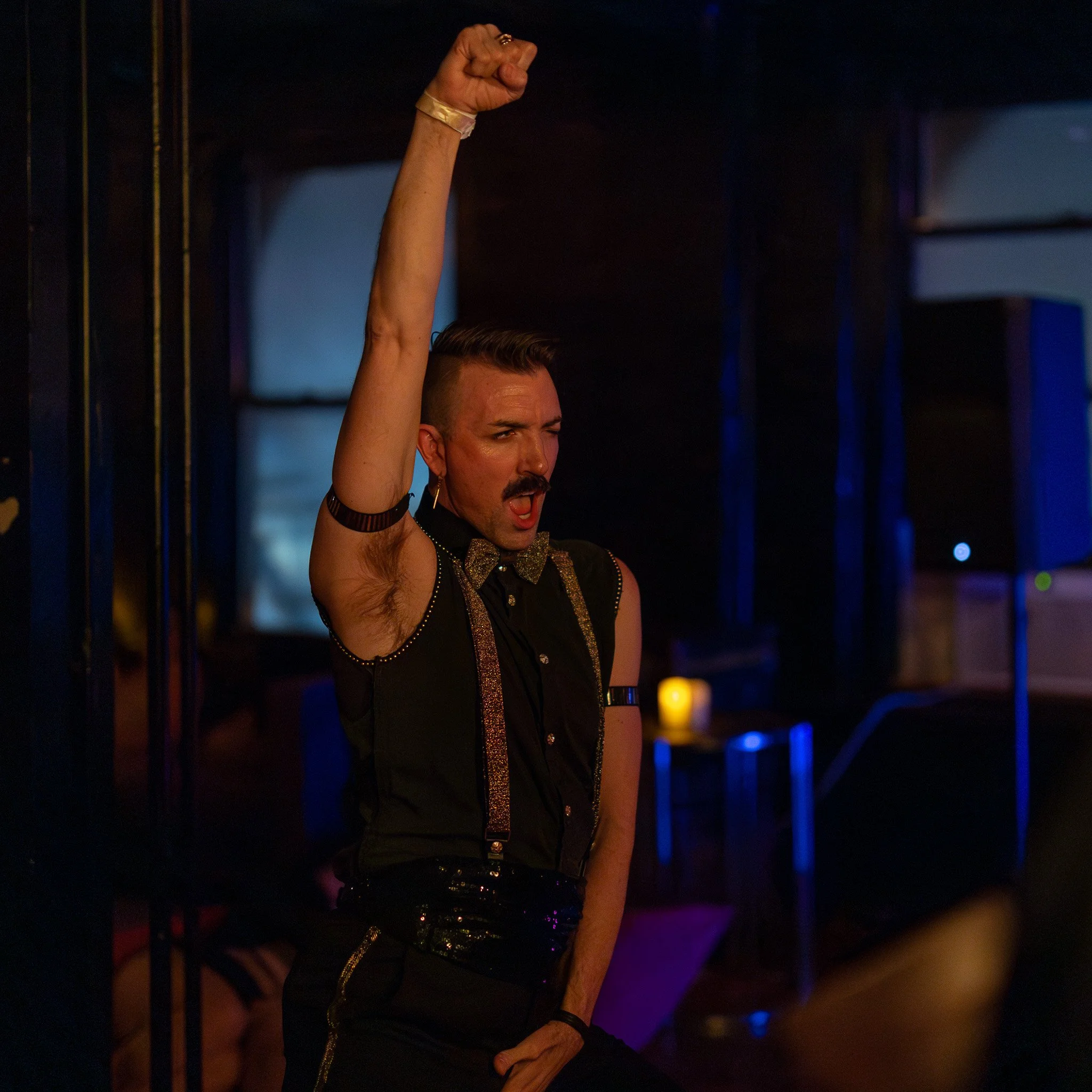 A man with a mustache, wearing a black shirt with suspenders and a gold bow tie, standing with his right fist raised and mouth open in a lively setting with dim lighting.