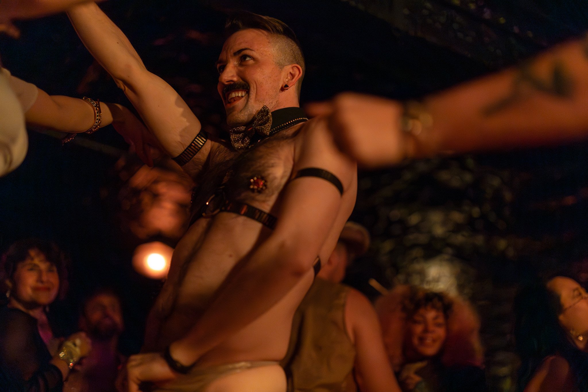 A man with a beard and tattoos, wearing a bow tie, is smiling while dancing closely with a person in a leotard, in a lively, dimly lit party or nightclub setting with other smiling people in the background.