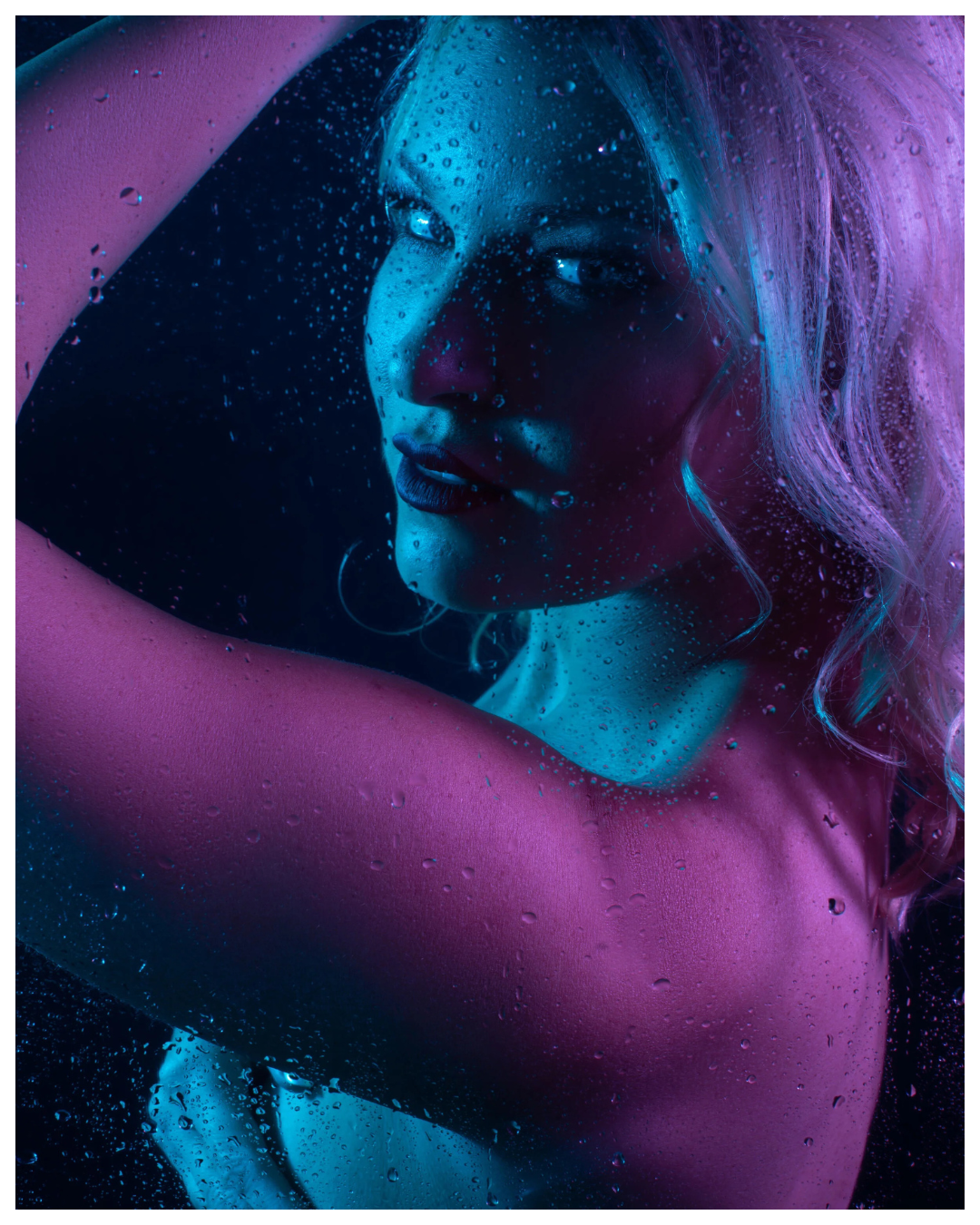 A woman with light blonde hair in neon pink and blue lighting, looking through a rain-covered glass surface.