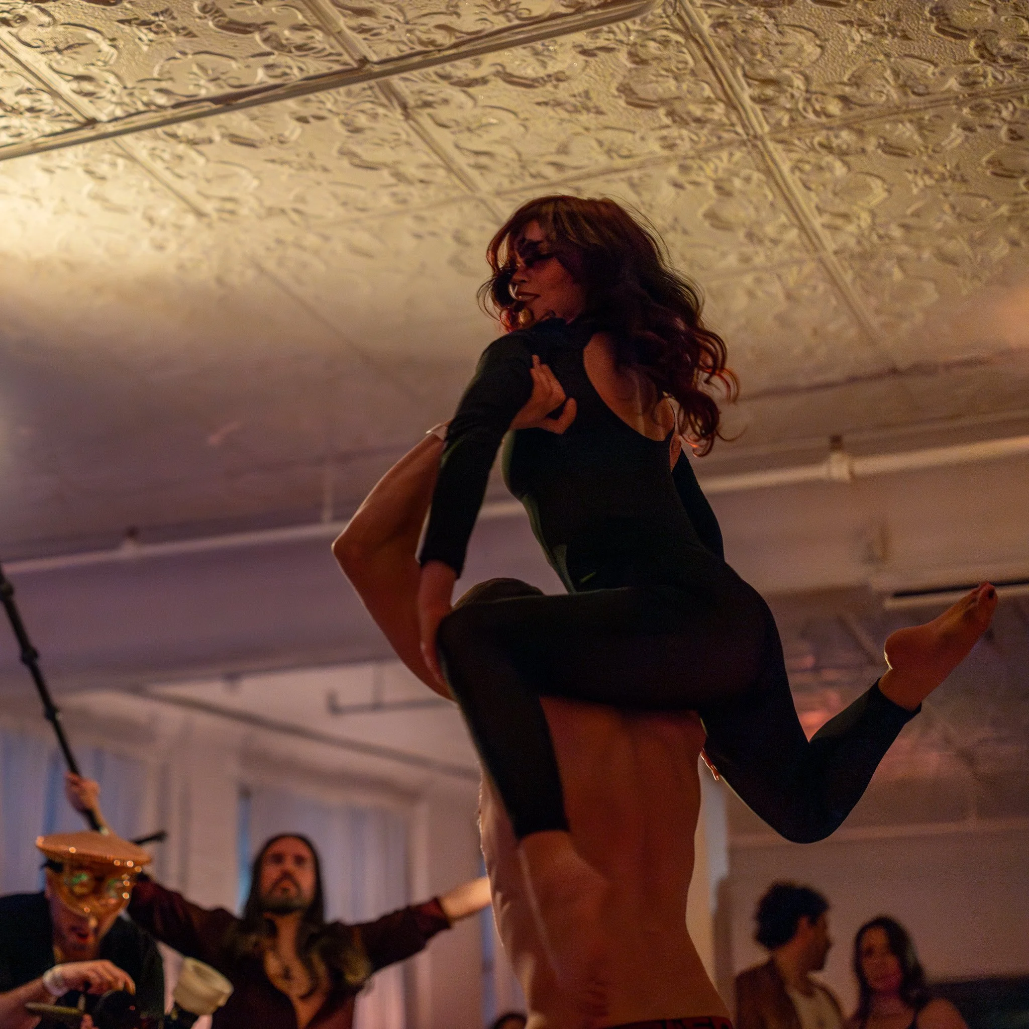 A woman in black dance clothing being lifted and spun in a dance performance, with a decorated ceiling and other spectators in the background.
