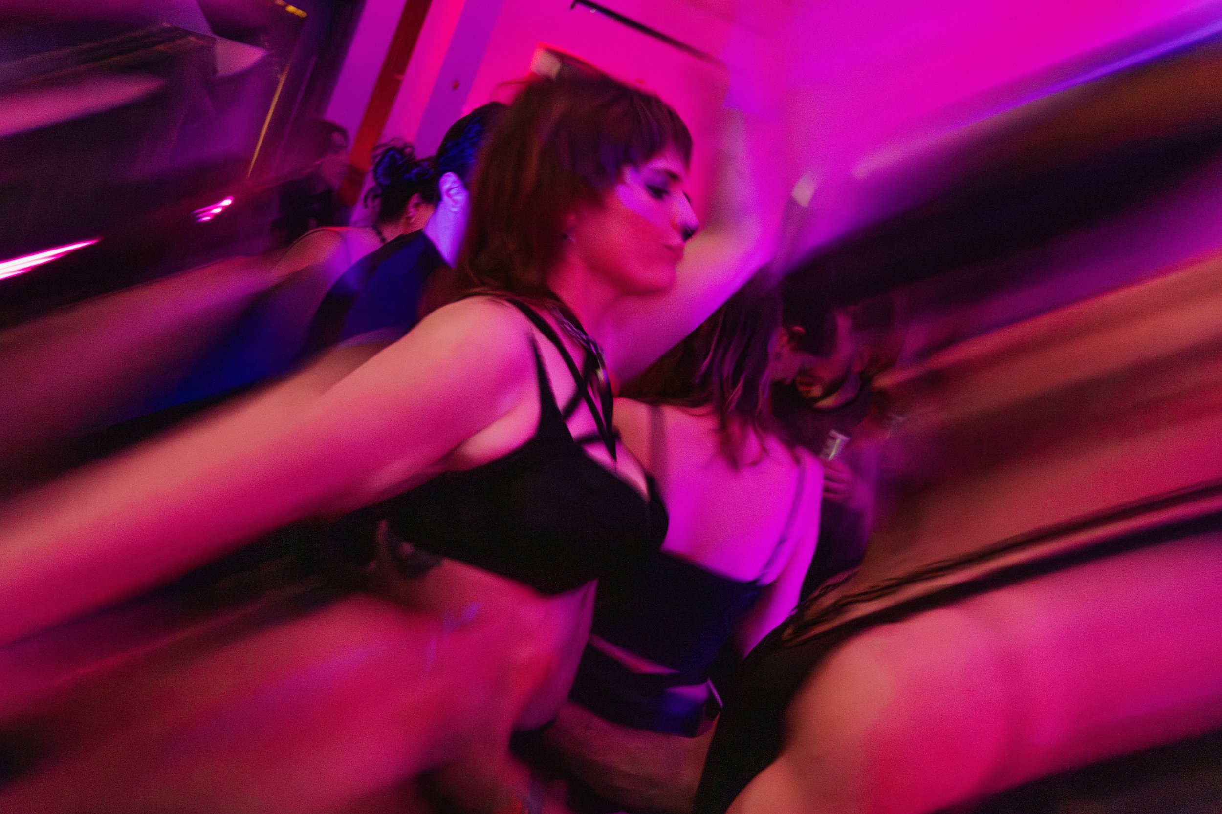 People dancing at a nightclub with colorful pink and purple lighting, some with their eyes closed and arms raised.