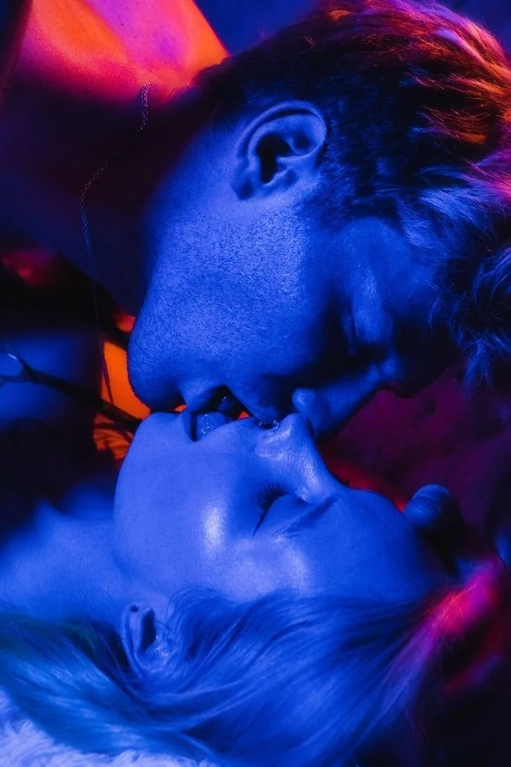 A close-up of two people with faces close together, almost kissing, illuminated by vibrant blue, pink, and orange neon lights.