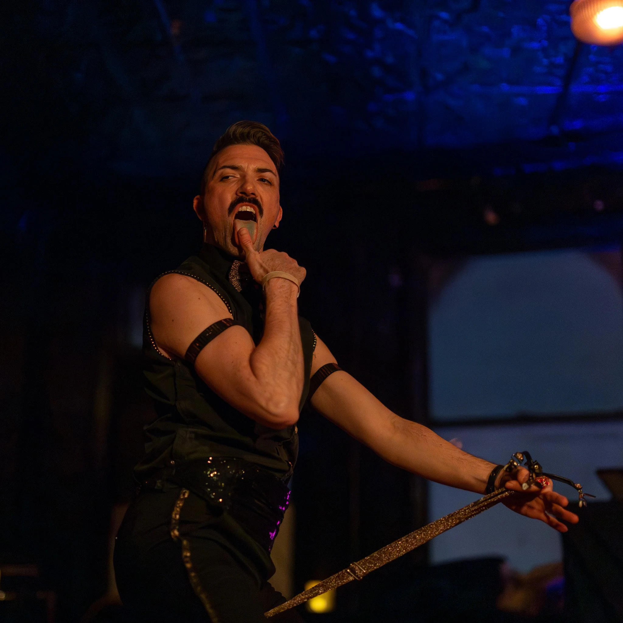 Performer on stage mid-performance, holding a glittery microphone stand, under dim stage lighting with a dark ceiling background.