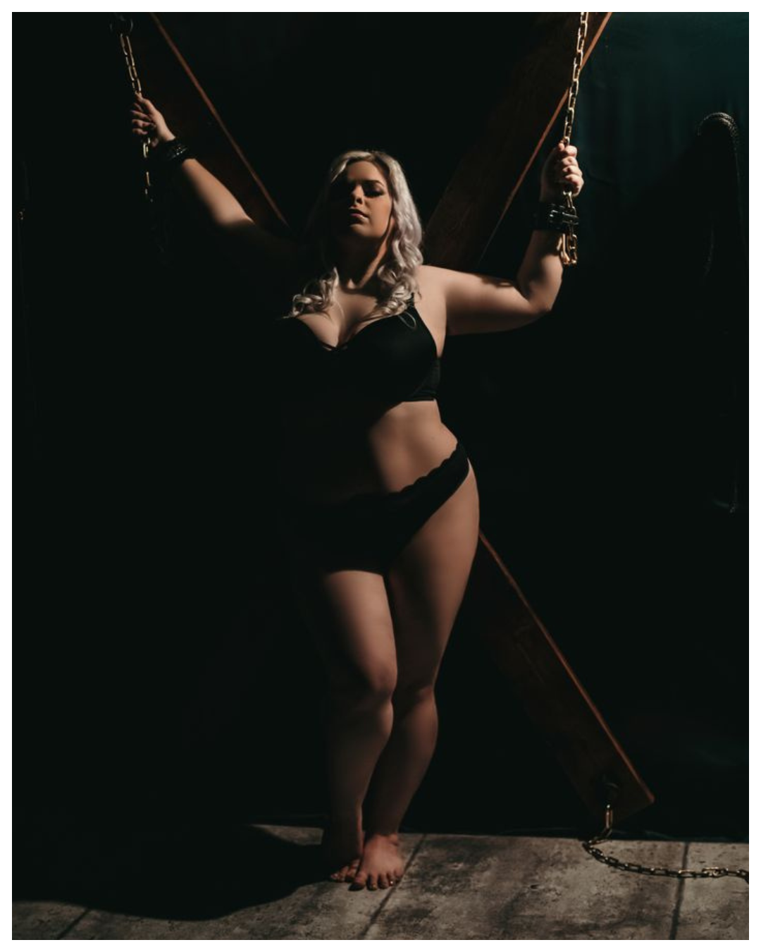 A woman with blonde curly hair in black lingerie, chained to a wooden cross with chains, standing barefoot on a dark floor in a dimly lit setting.