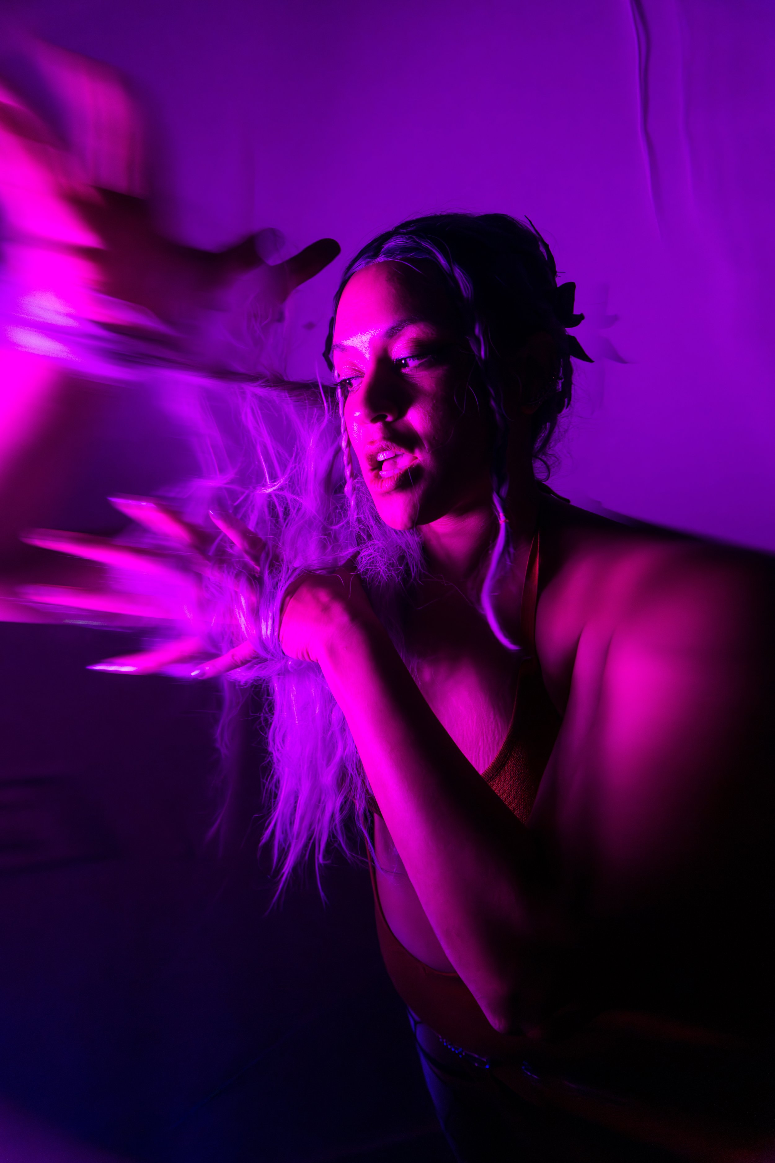 Person with long, curly hair illuminated by purple and pink lighting, striking a dynamic pose with long hands and an expressive face.
