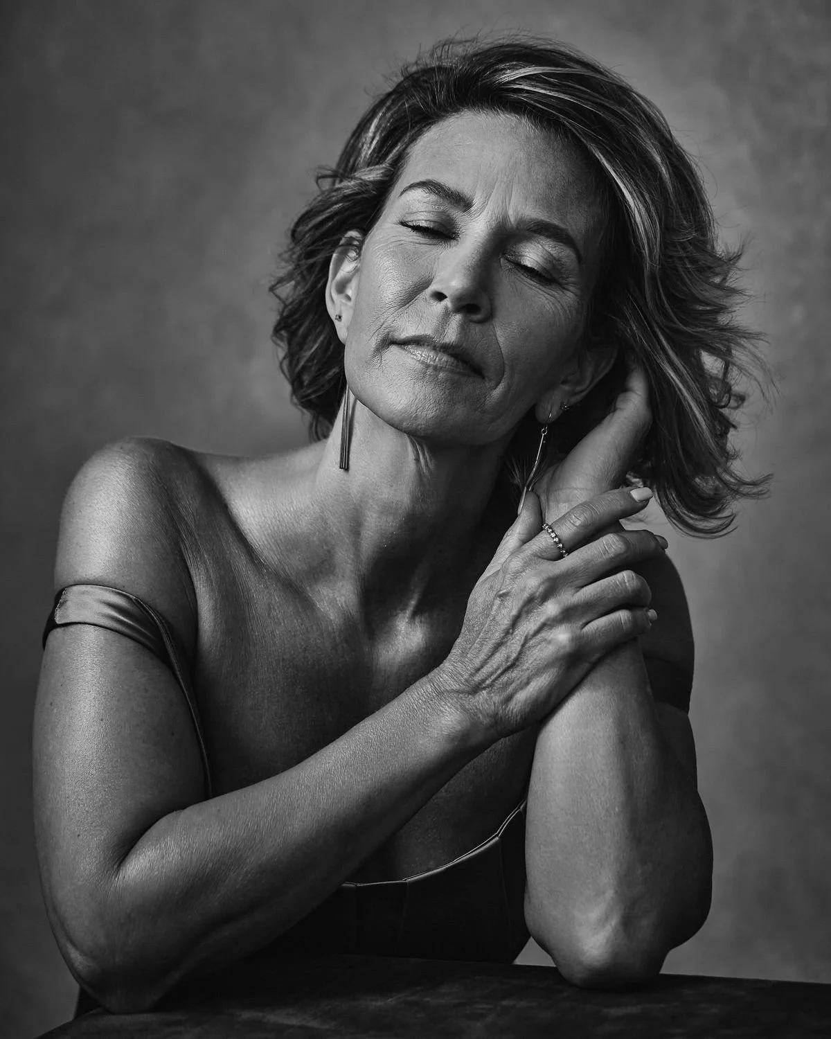 Black and white portrait of a middle-aged woman with short wavy hair, closed eyes, and a serene expression, touching her face with one hand and resting the other on a surface.