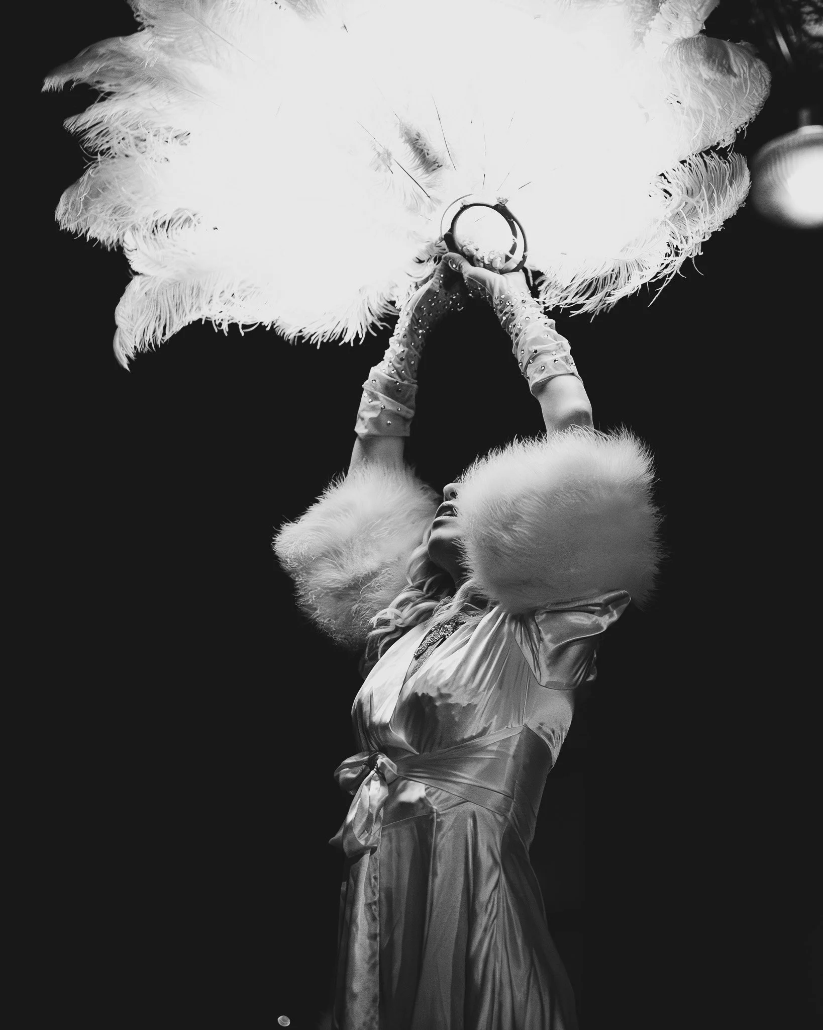 A performer in a shiny dress and fur hat, holding a large feathered fan above her head, possibly on stage during a performance.