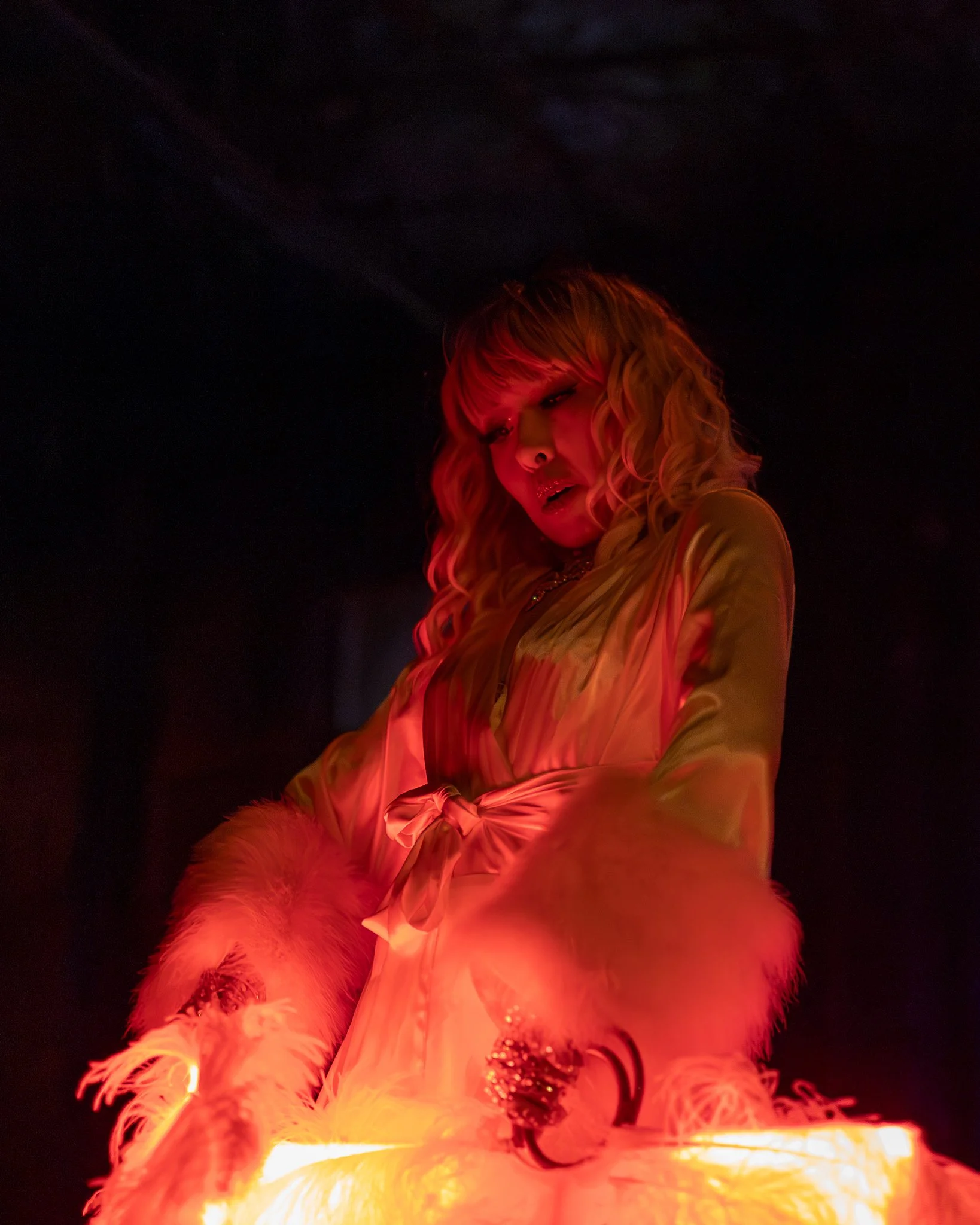 A woman with curly blonde hair, wearing a satin robe with fur trim, is looking down with a surprised or intense expression on her face, illuminated by a warm glow in a dark setting.