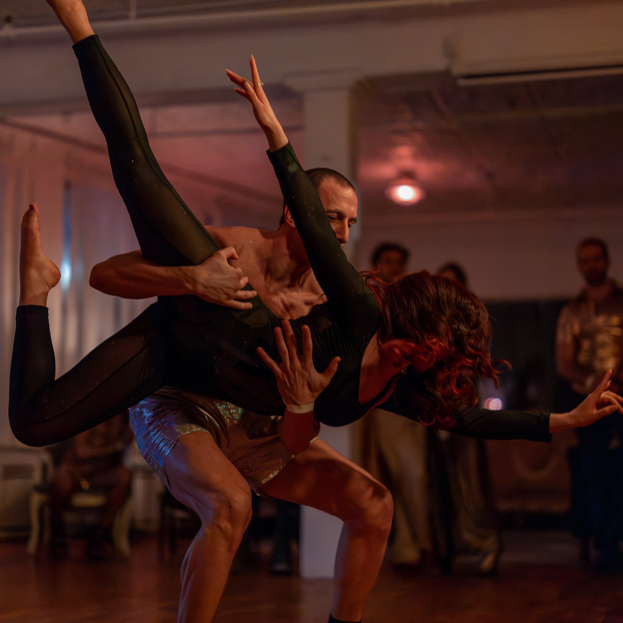 A man lifting a woman in a dance or acrobatic pose during a performance, with onlookers in the background.