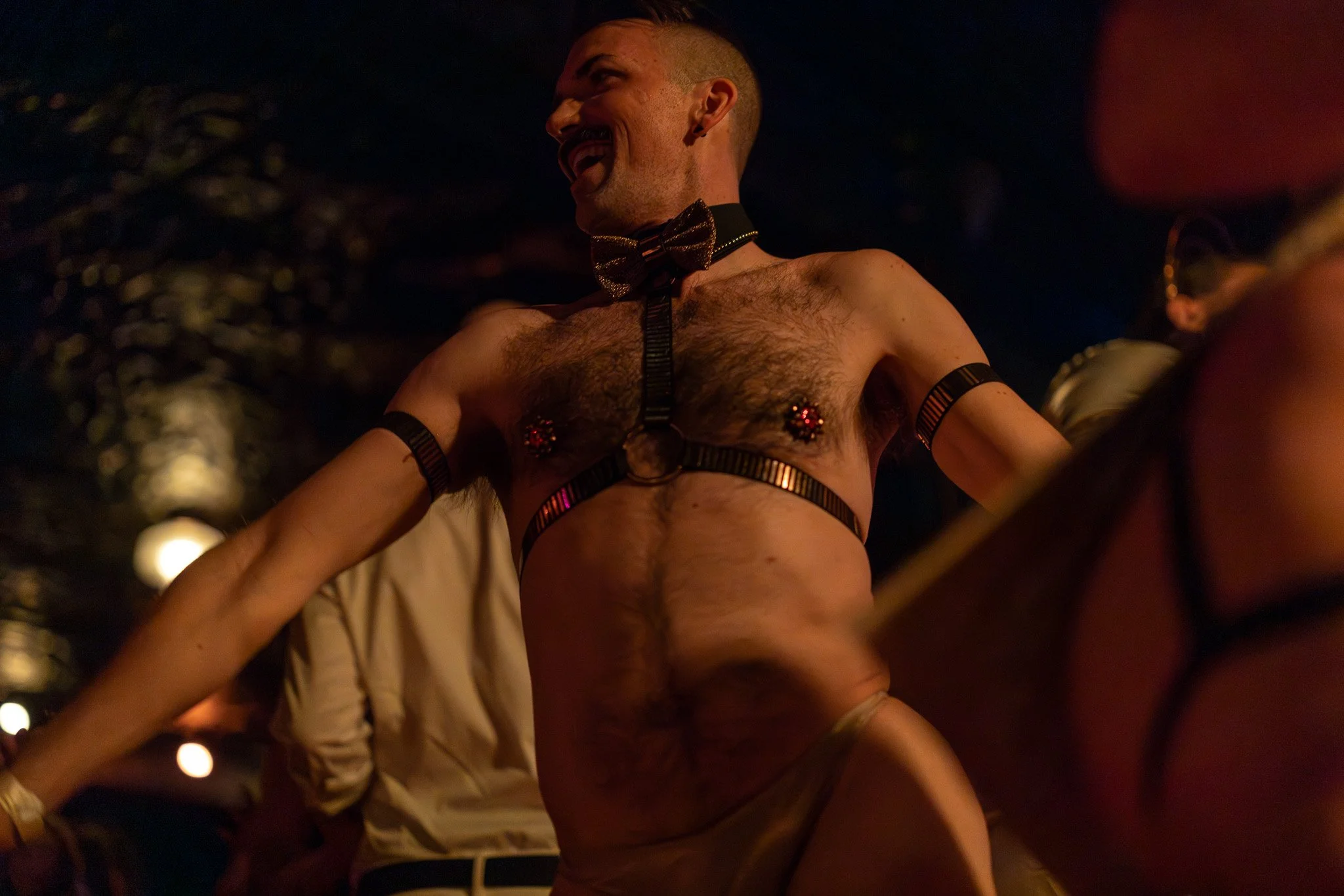 A person with a mustache and shaved head, wearing a bowtie, harness, and nipple decorations, dancing or performing in a dimly lit setting.