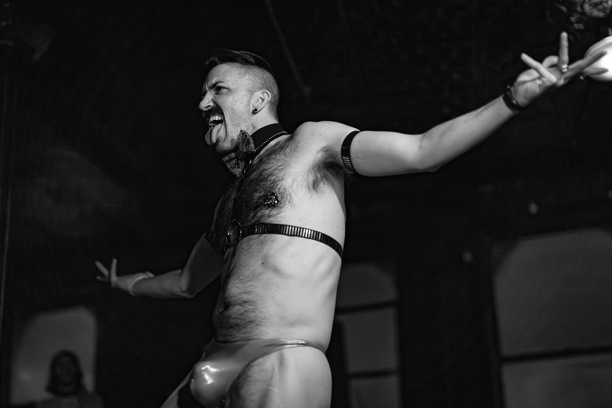 A man with short hair, an earring, and a bow tie peeks out from behind a wall, sticking out his tongue and making a funny face, while posing with one arm extended.