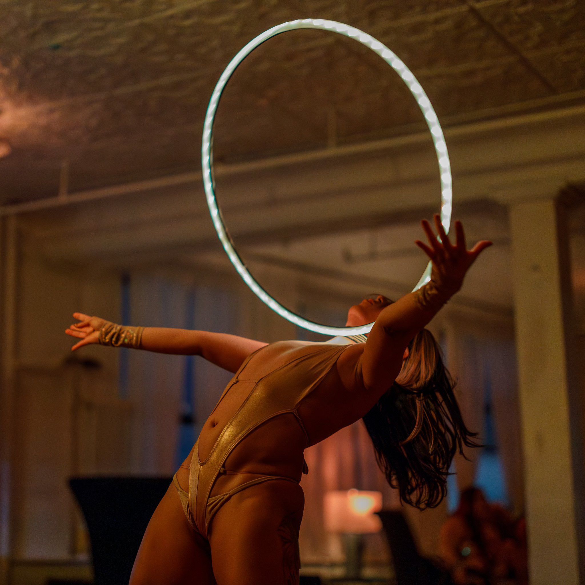 A female performer in gold outfit performing with a hoop in a dimly lit room.