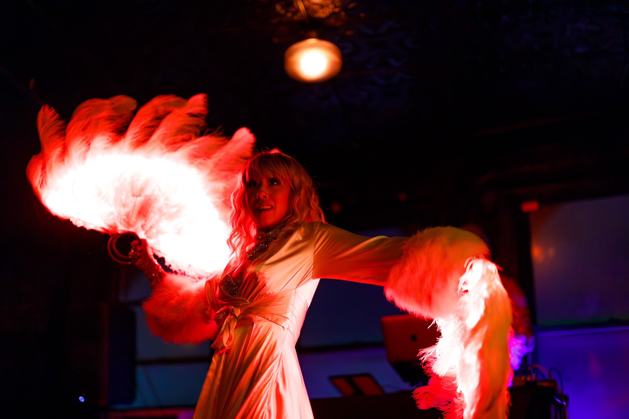 A woman performing a fire dance, holding a flaming torch with large feathers attached, in a dimly lit setting.
