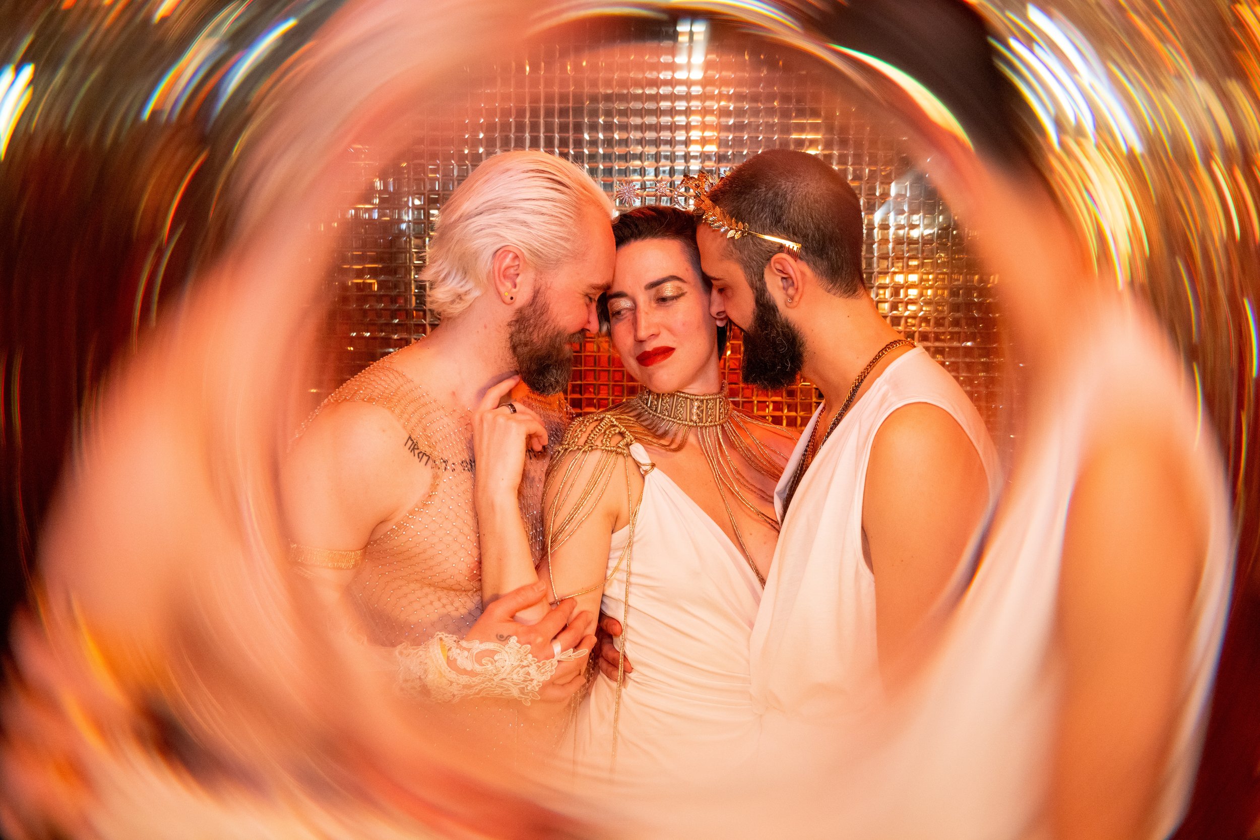Three individuals in elaborate costumes hugging and touching foreheads, surrounded by a swirling frame with a shimmering, golden background.