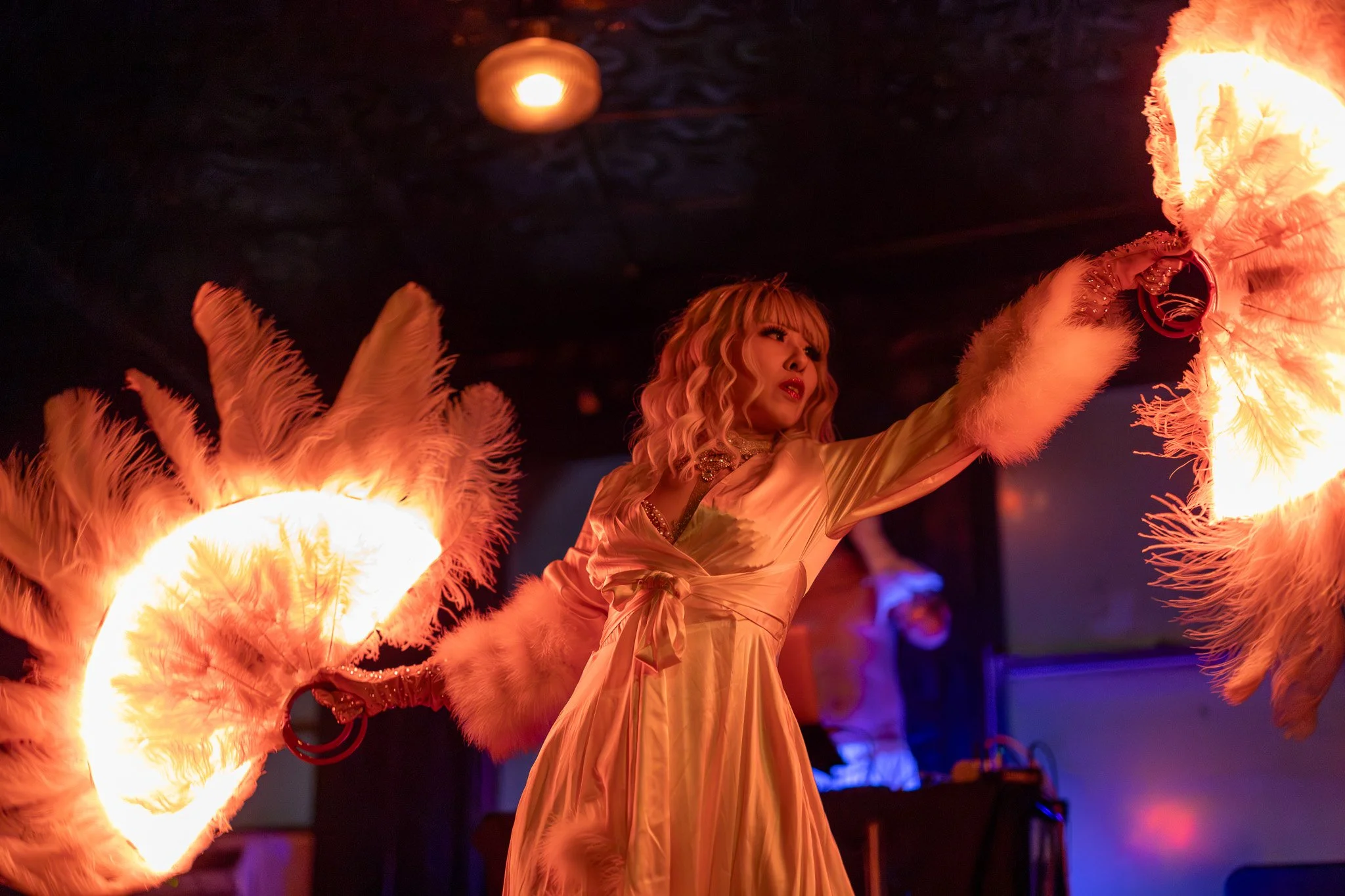 A female performer dressed in a satin gown with fluffy cuffs is performing a fire dance, holding lit fire fans with feathers illuminated by the flames in a dimly lit room.