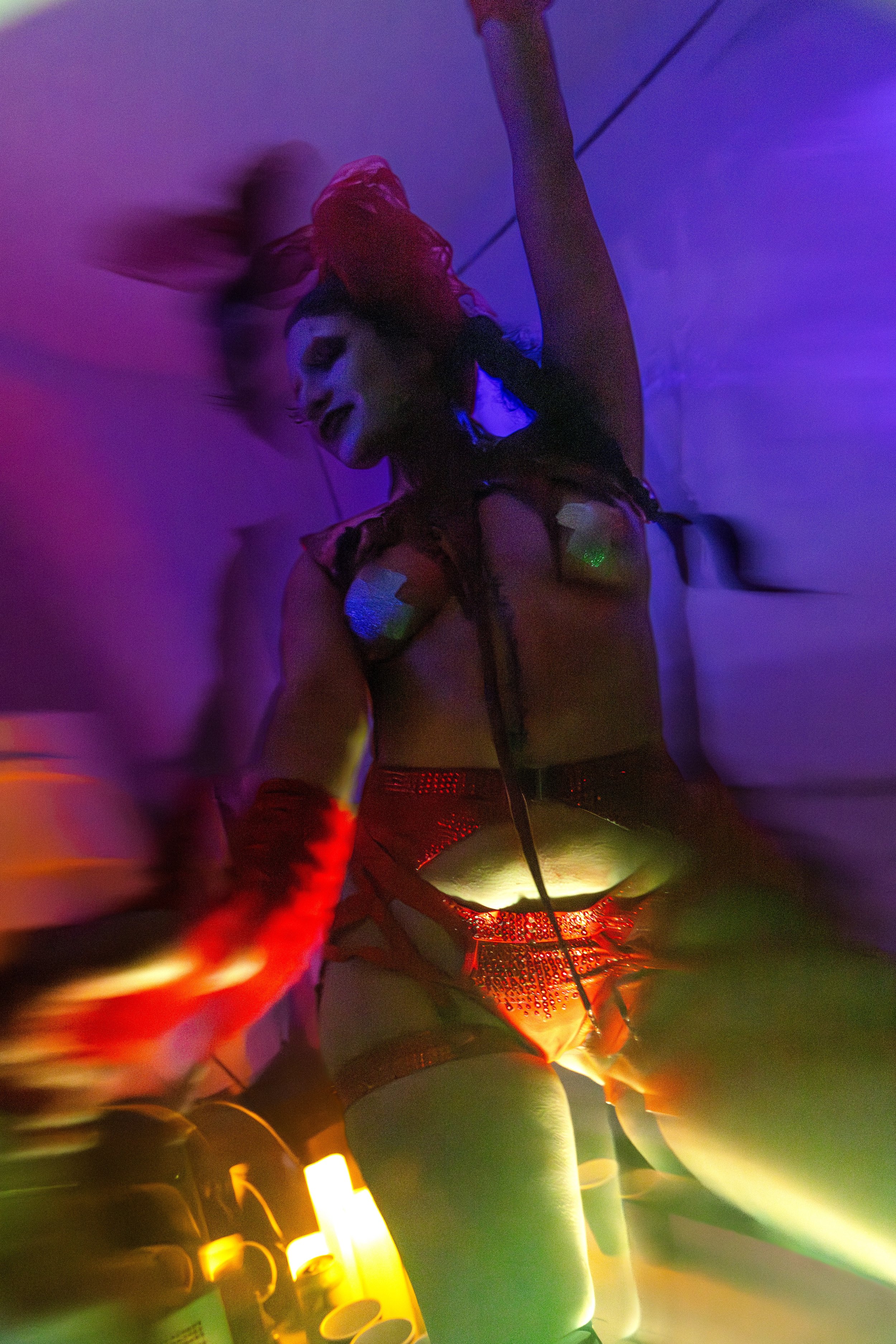 A performer with red hair and makeup, wearing glittery attire and red gloves, dances with arms raised in a colorful, neon-lit setting.