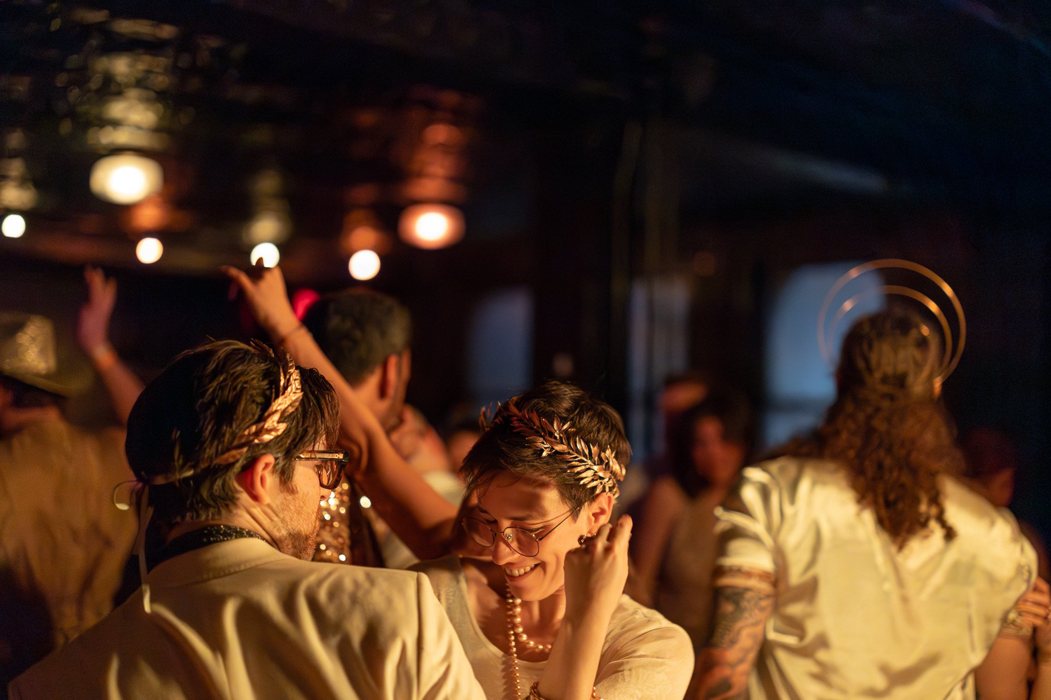 People dancing at a party decorated with string lights, with some wearing laurel wreath crowns.