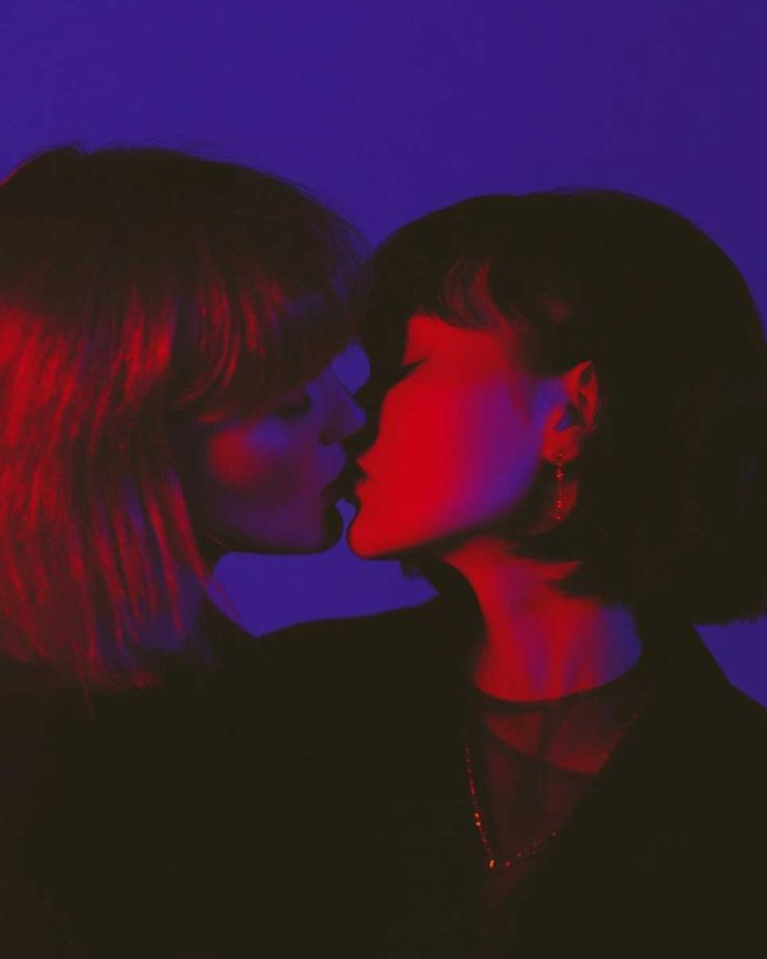 Two women with dark hair and earrings are close to each other, appearing about to kiss or in an intimate moment, illuminated by red, purple, and blue lighting.