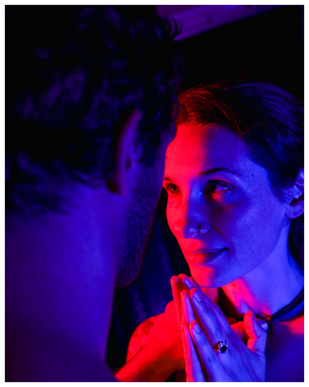 A woman with dark hair and a nose ring gazes into a man's eyes, her hands pressed together near her face, illuminated by red and blue lighting.