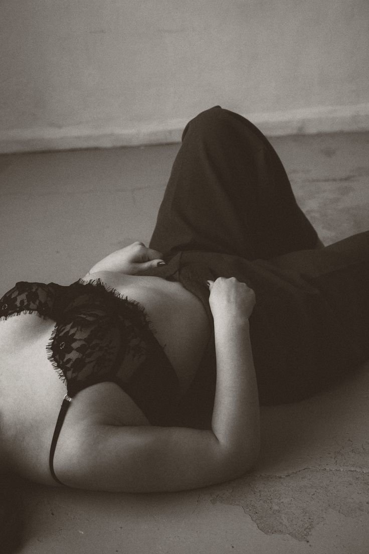 A person lying on the floor, dressed in dark clothing including a lace top, with one arm resting on their stomach and their hand near their chest.