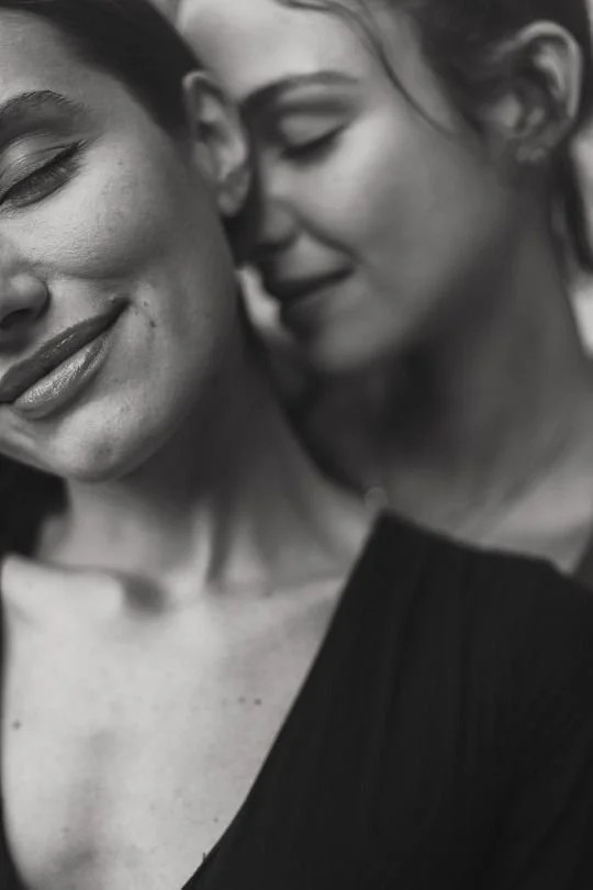 A close-up black and white photo of two women smiling and leaning close to each other with eyes closed.