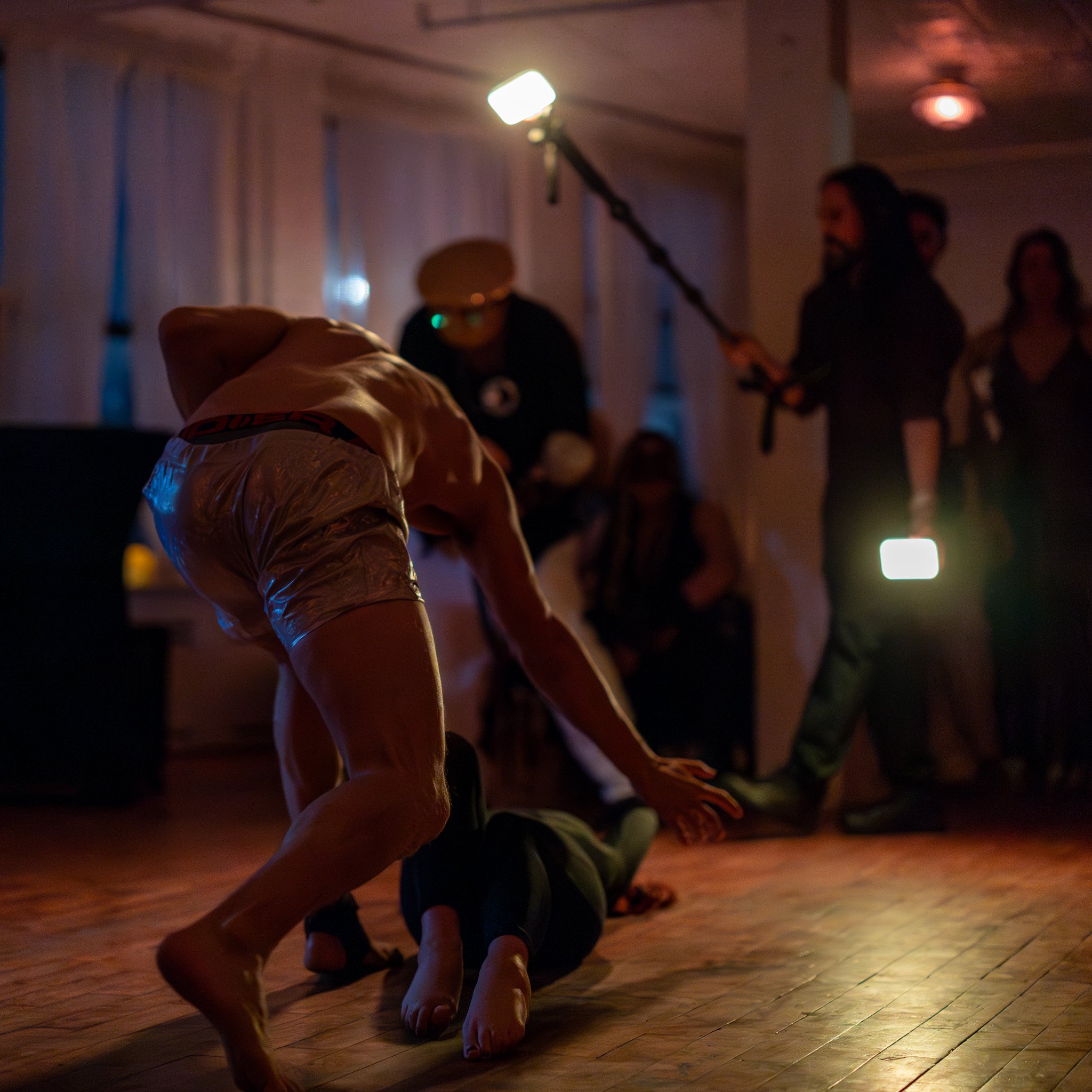 A scene on a dimly lit dance floor showing a person on the ground with another person leaning over him, possibly in a fighting or wrestling pose. In the background, there are people, one holding a boom microphone and another holding a light, indicati